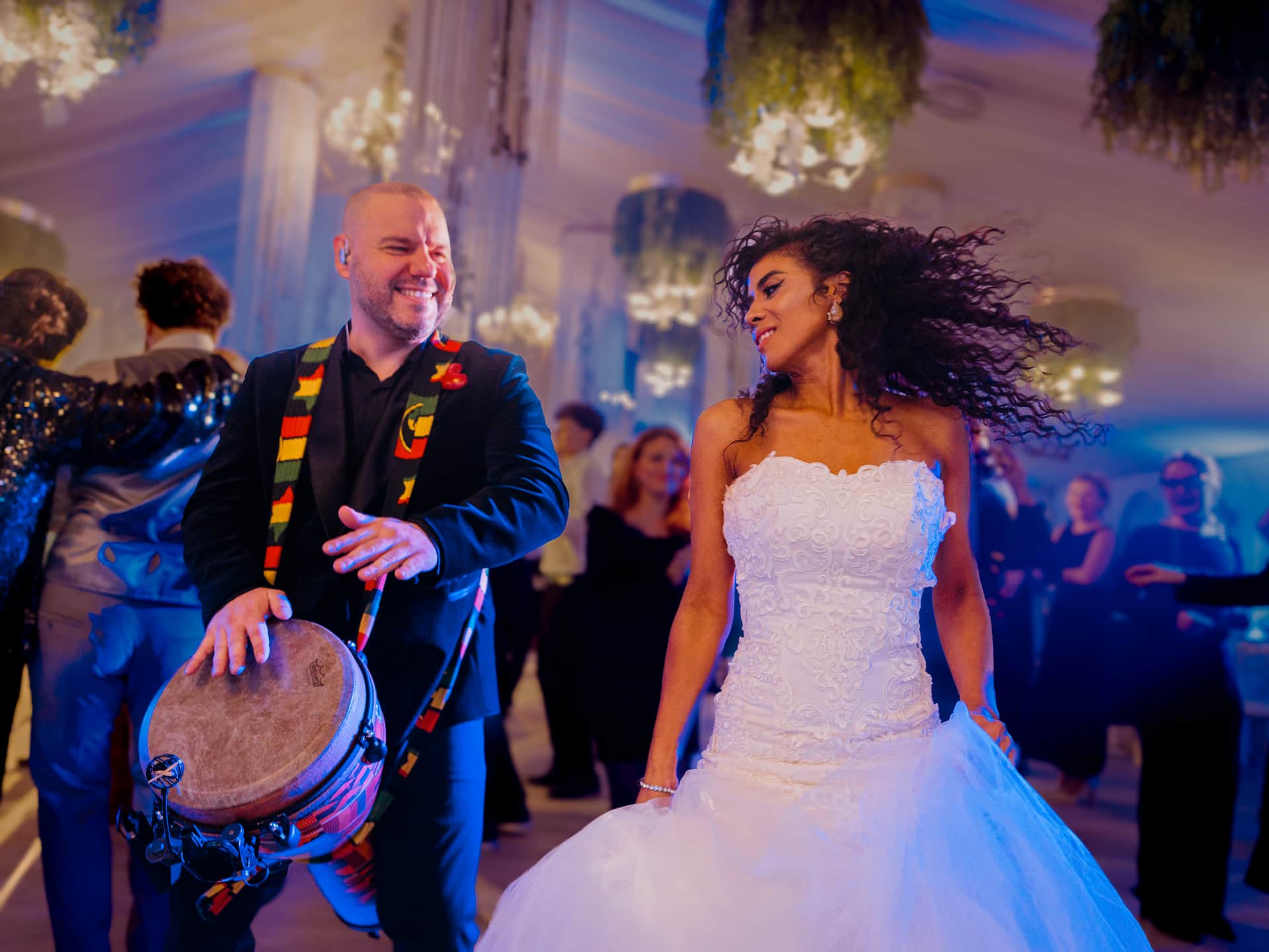 Bărbat zâmbind cântând la toba djembe lângă o femeie care dansează în rochie albă de mireasă la o petrecere elegantă.