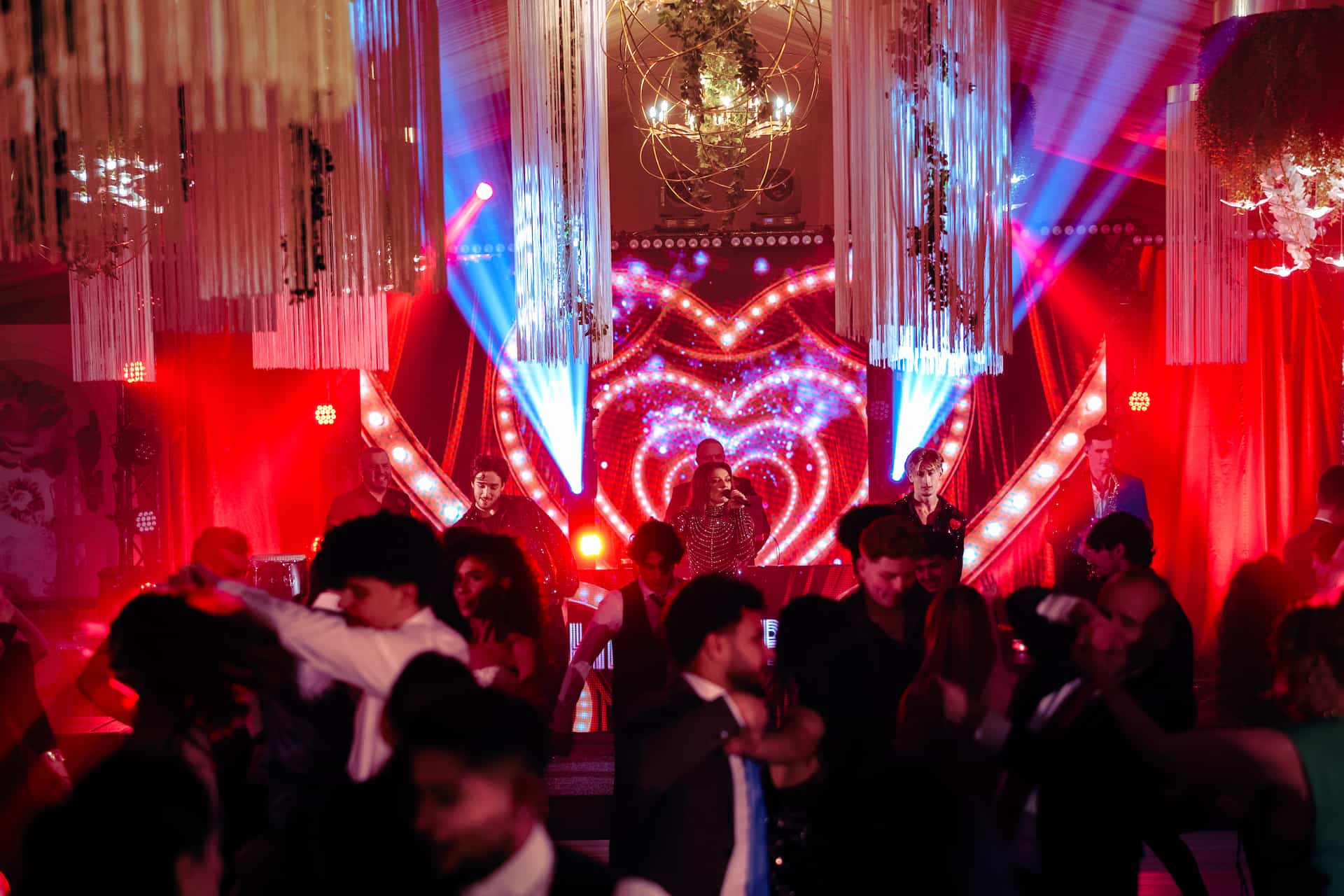 O trupă cântă pe scenă cu un fundal luminos în formă de inimă, iar oameni dansează în față sub lumini roșii și albastre.
