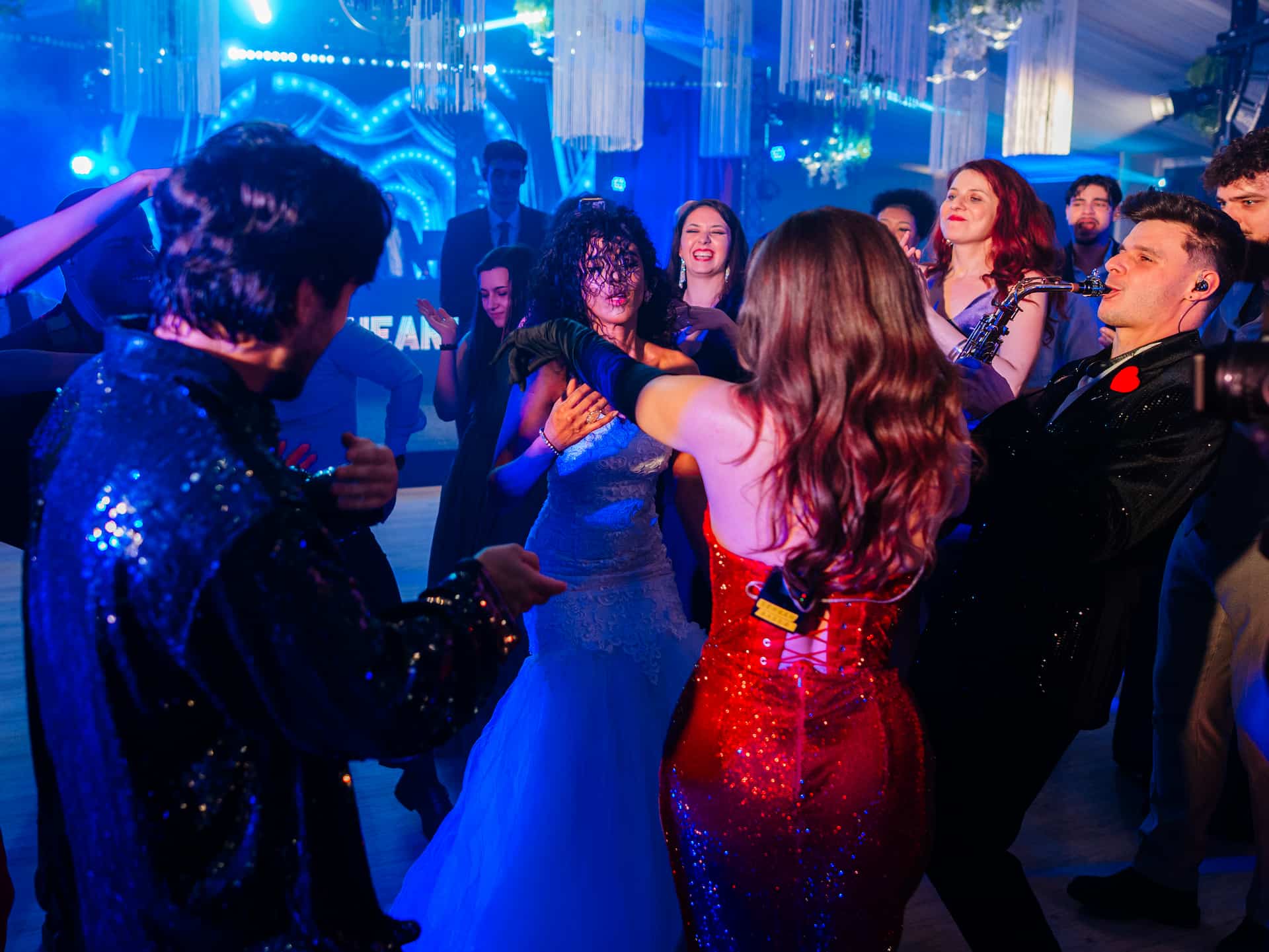 O petrecere în interior cu oameni dansând sub lumini albastre și albastre-albastre, o femeie în rochie de mireasă și un muzician cântând la saxofon.