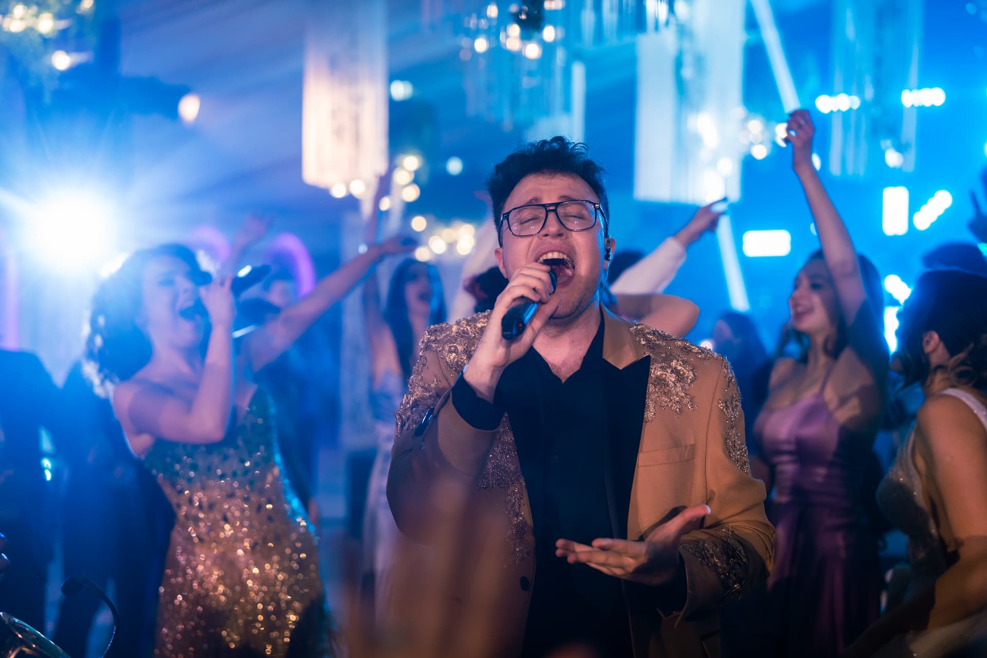 Un bărbat cu ochelari cântă la microfon într-un club plin de oameni care dansează și se distrează sub luminile albastre.