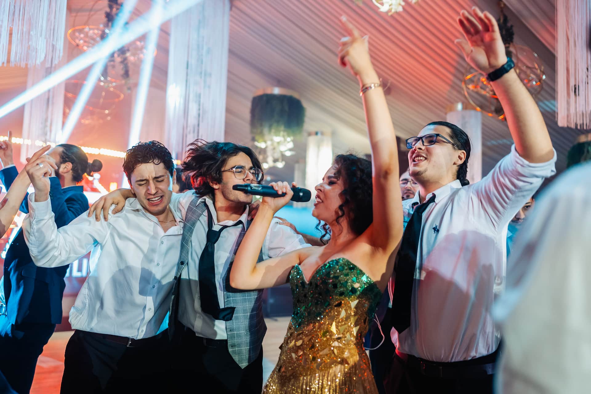 Tineri la o petrecere, cântând la microfon și dansând cu brațele ridicate într-un decor luminat festiv.