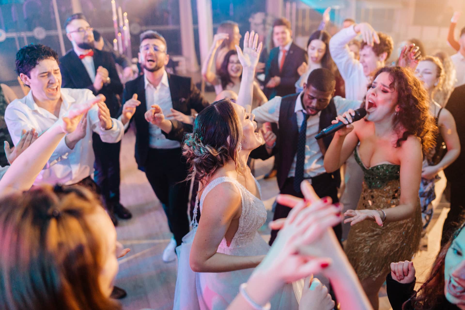 O mireasă și o femeie cântând la microfon în timp ce oamenii din jur dansează și se distrează la o petrecere.