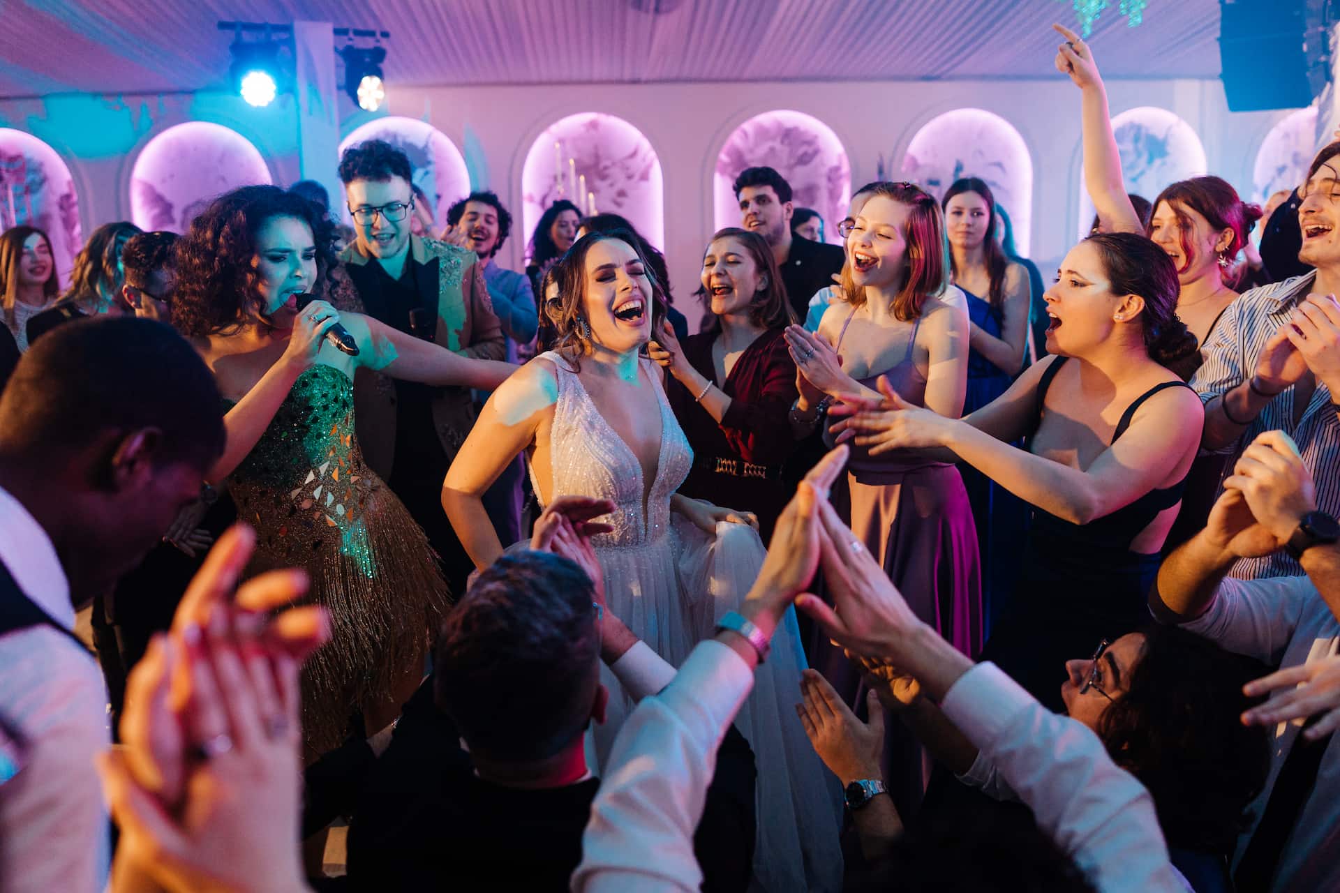 O mireasă zâmbitoare dansează și cântă înconjurată de invitați veseli care o aplaudă și o încurajează la o petrecere.