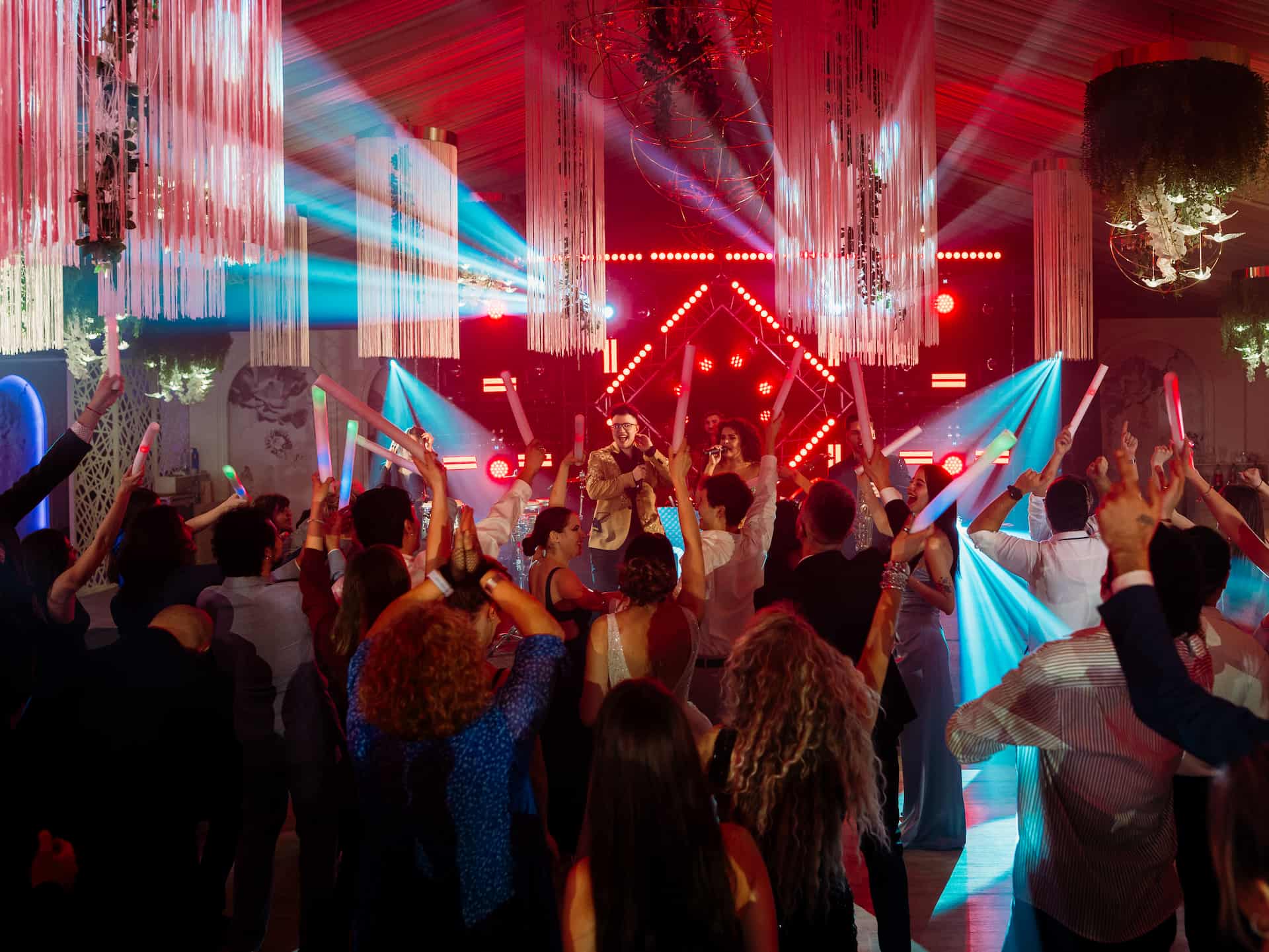 O mulțime de oameni la un concert sau petrecere, ținând baghete luminoase, cu doi cântăreți pe scenă și lumini spectaculoase roșii și albastre.