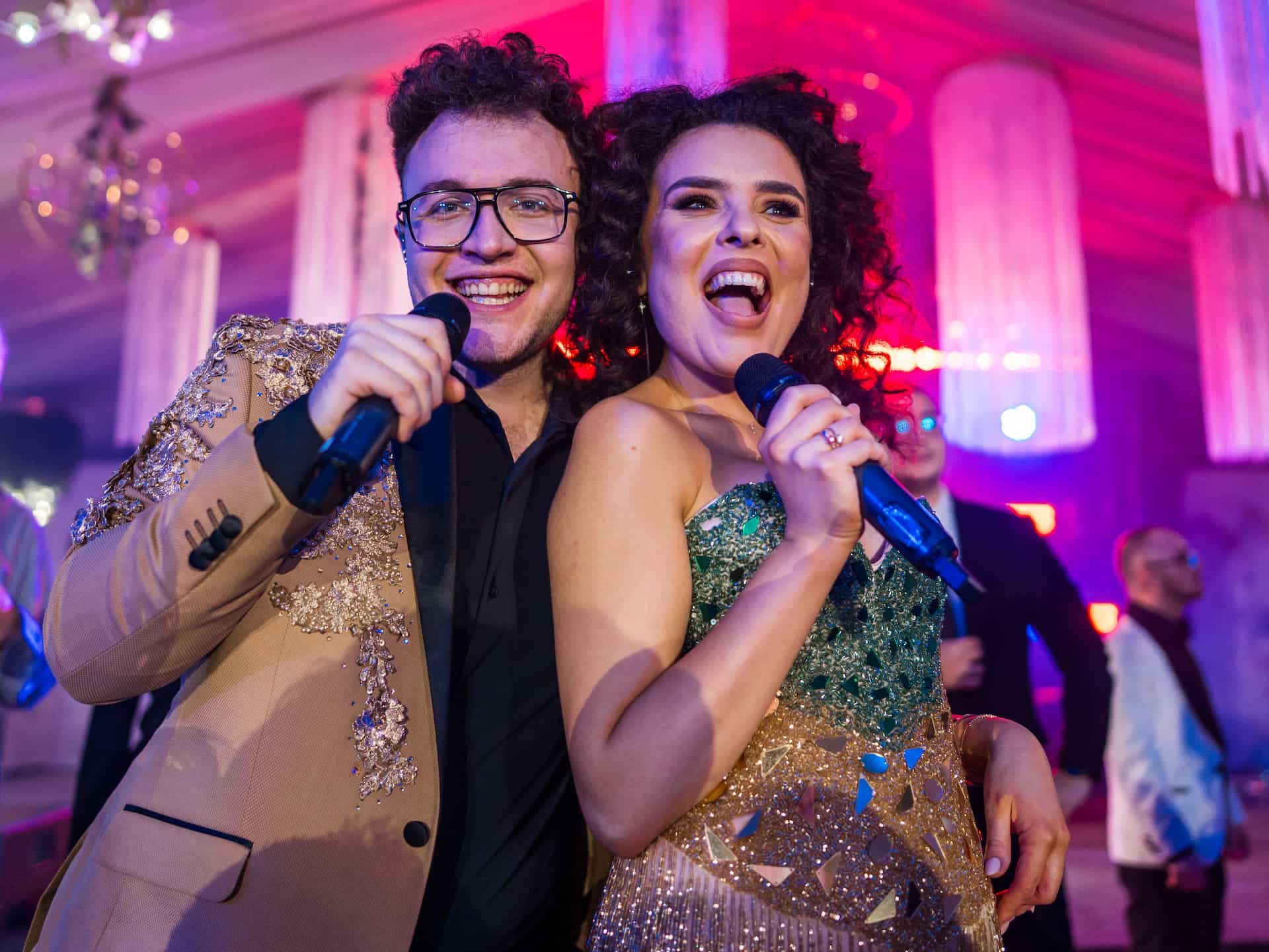 Bărbat și femeie în haine elegante cântând cu microfoane într-un cadru festiv cu lumini colorate.