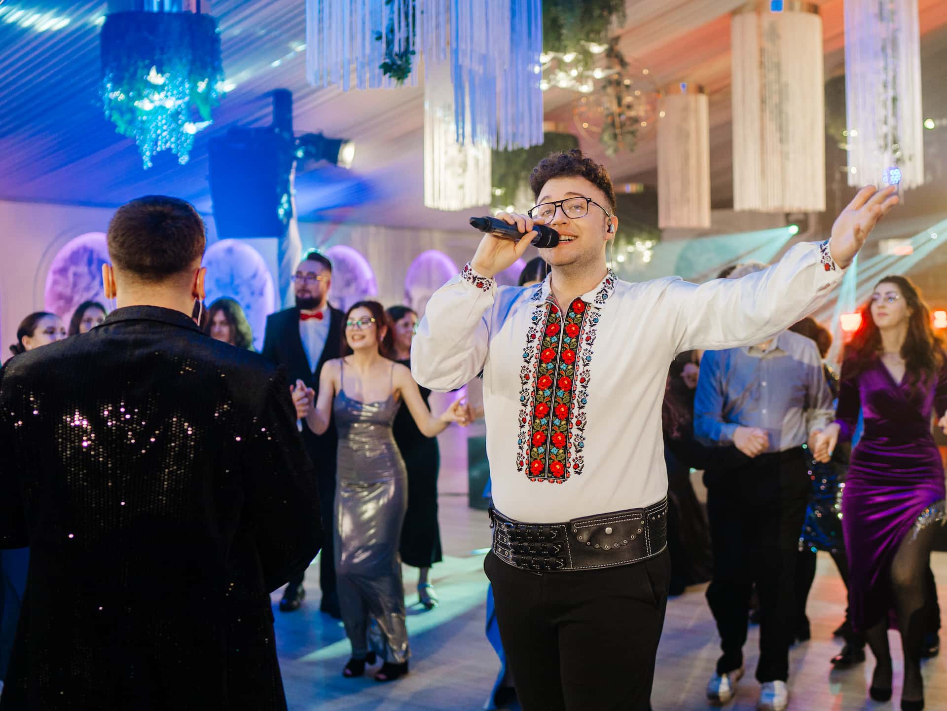 Bărbat care cântă la microfon îmbrăcat într-o cămașă tradițională cu broderie, înconjurat de oameni care dansează la o petrecere elegantă.