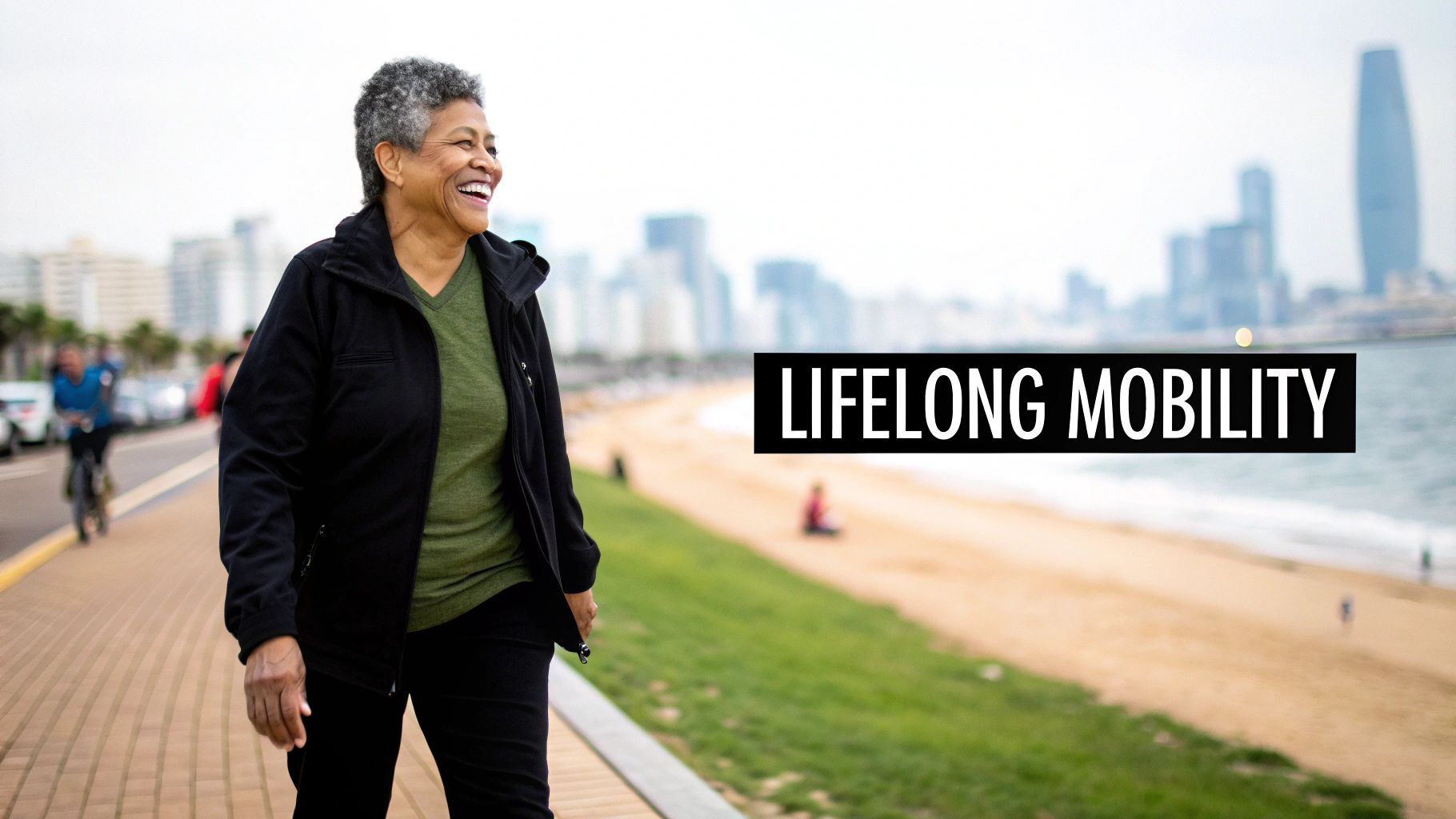 Happy senior woman in a black jacket and green shirt walks by a beach with a city skyline.
