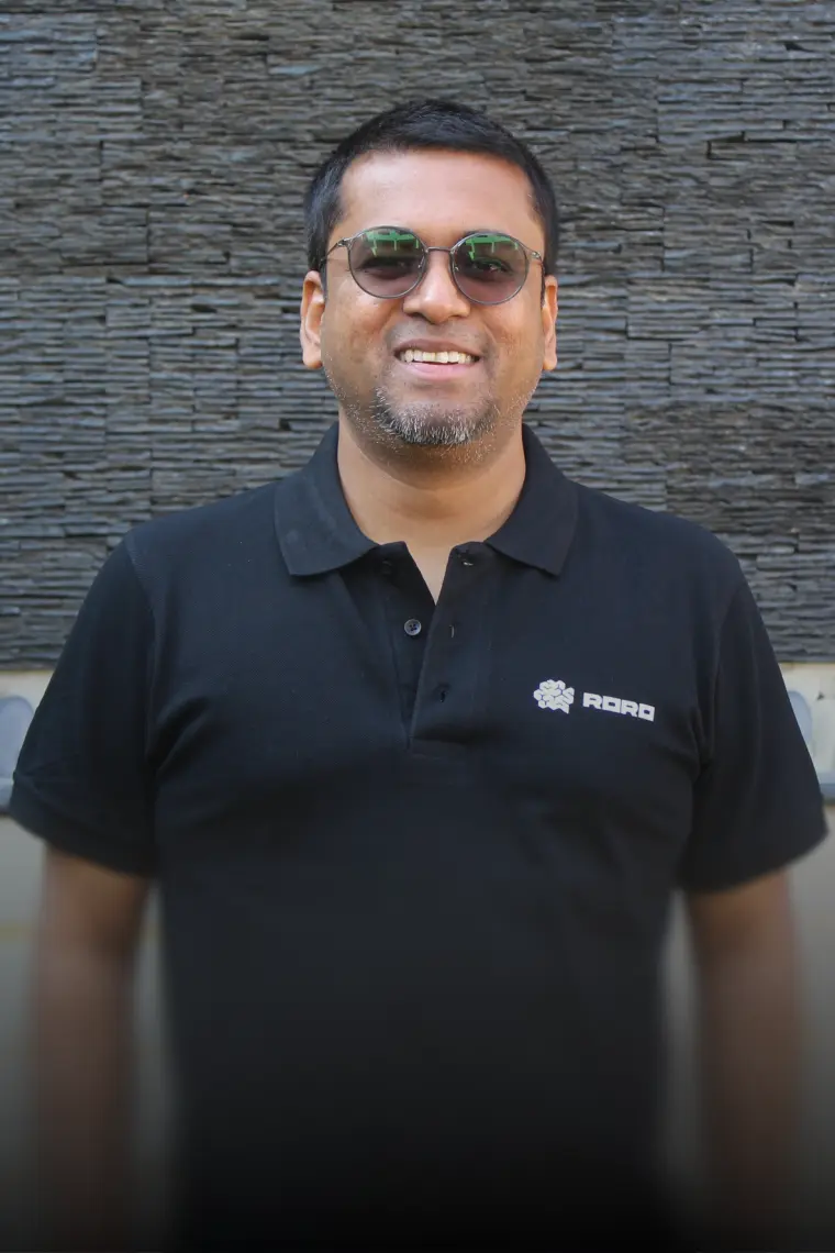 Achin wearing sunglasses and a black polo shirt with the RORO logo, smiling in front of a textured dark stone wall.
