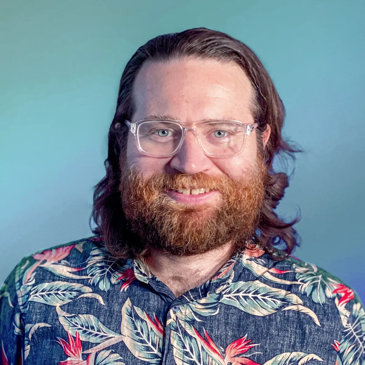 Van Tucker from LuxorOne with a full beard, long hair, clear glasses, and a blue floral shirt against a plain blue background.