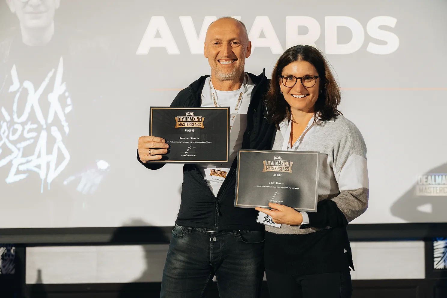 Zwei lächelnde Personen halten Urkunden der Dealmaking Masterclass, stehend vor einer Wand mit dem Schriftzug AWARDS.