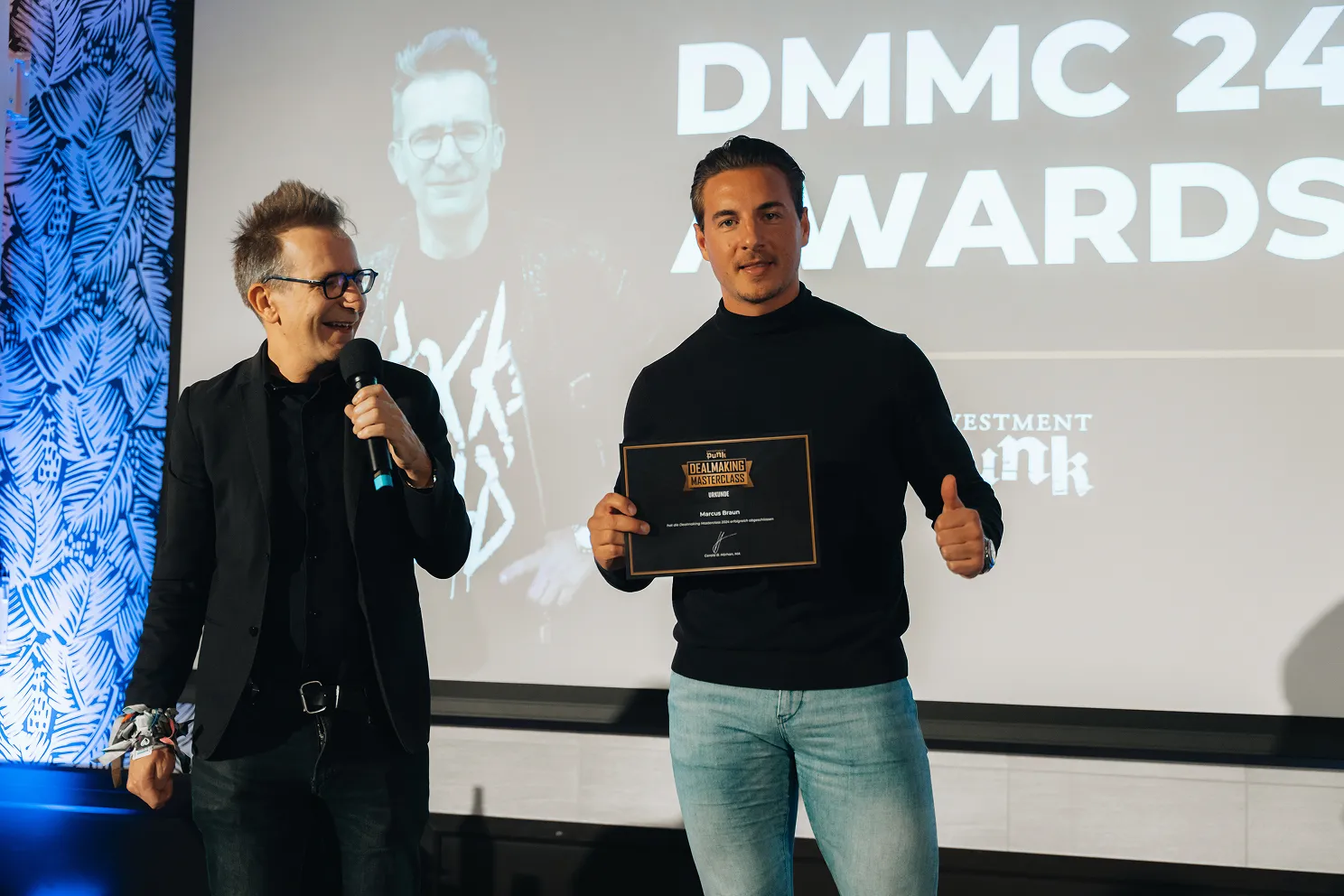 Zwei Männer bei der DMMC 24 Awards-Verleihung, der rechte hält eine Urkunde und zeigt Daumen hoch, der linke spricht in ein Mikrofon.
