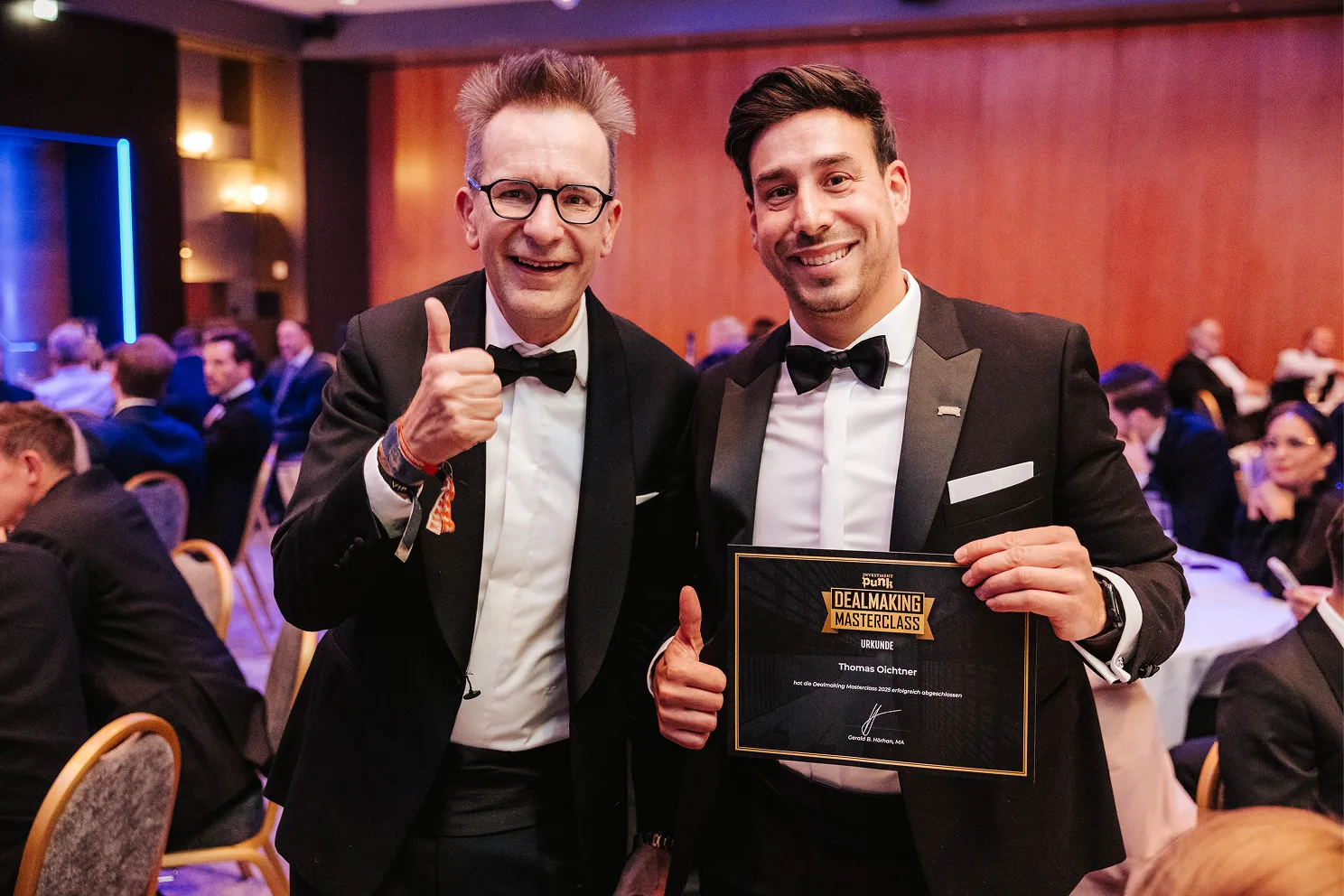 Zwei Männer in Anzügen und Fliegen, einer hält eine Urkunde der Dealmaking Masterclass und beide zeigen Daumen hoch.