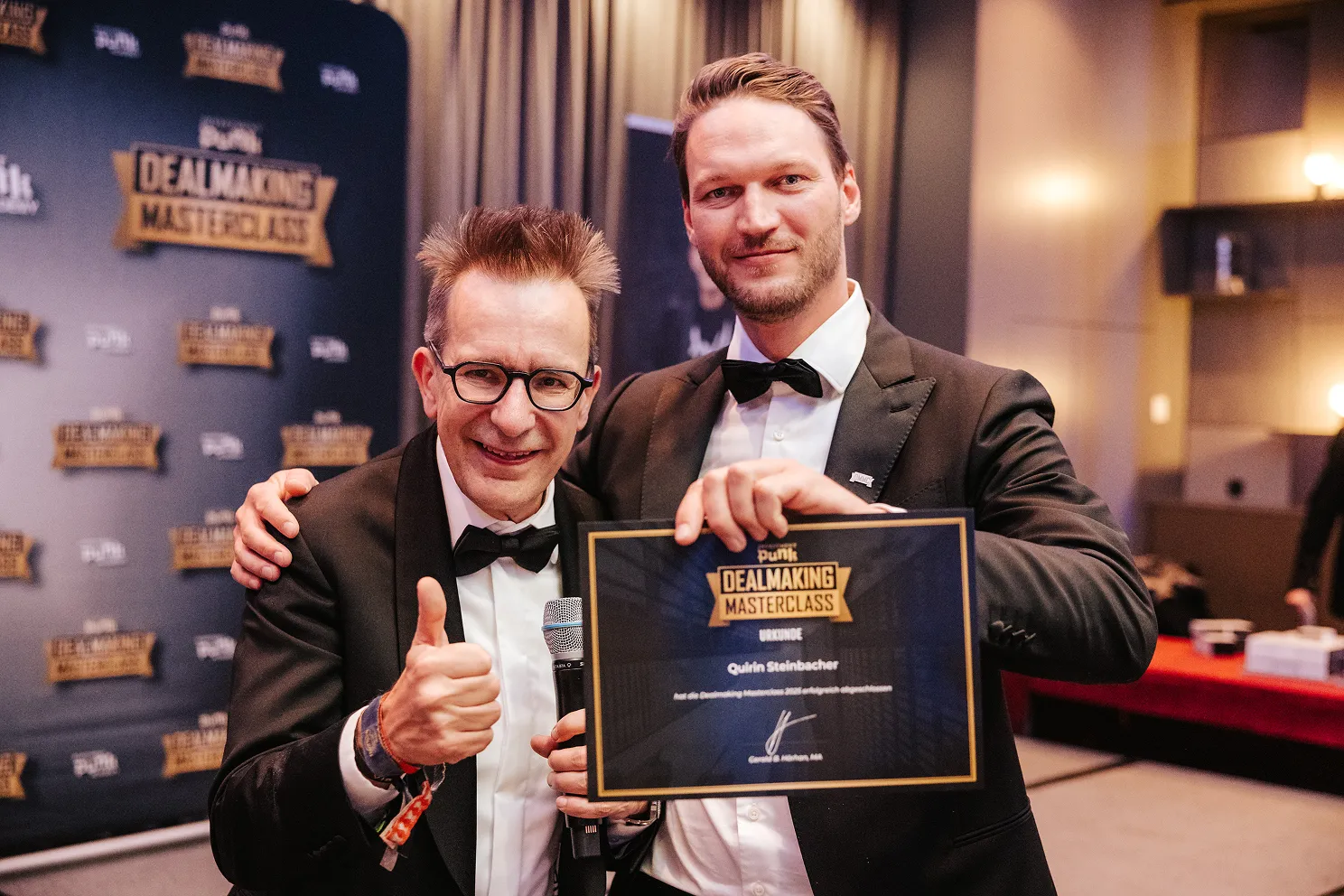 Zwei Männer in schwarzen Anzügen, einer hält eine Auszeichnung der DealMaking Masterclass, der andere zeigt einen Daumen hoch.