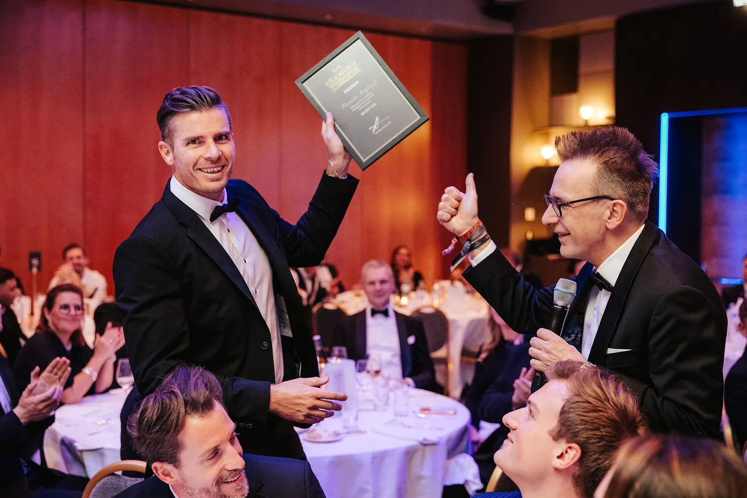 Mann in schwarzem Anzug hält einen Award in die Luft, während ein anderer Mann mit Mikrofon ihm zustimmend den Daumen hebt, Publikum applaudiert im Hintergrund.