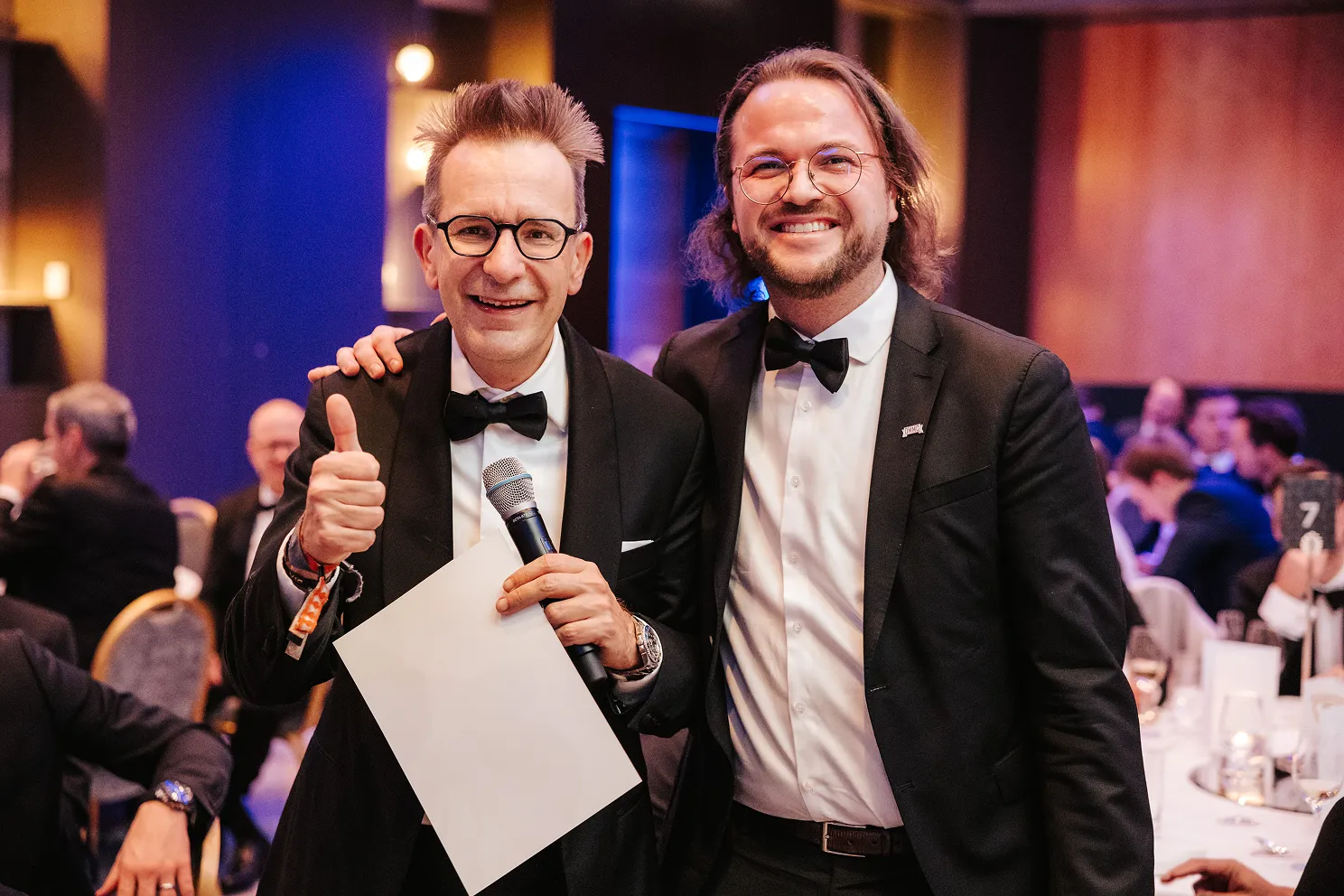 Zwei Männer in schwarzen Anzügen und Fliegen mit Mikrofon und Papier lächeln bei einer festlichen Veranstaltung.