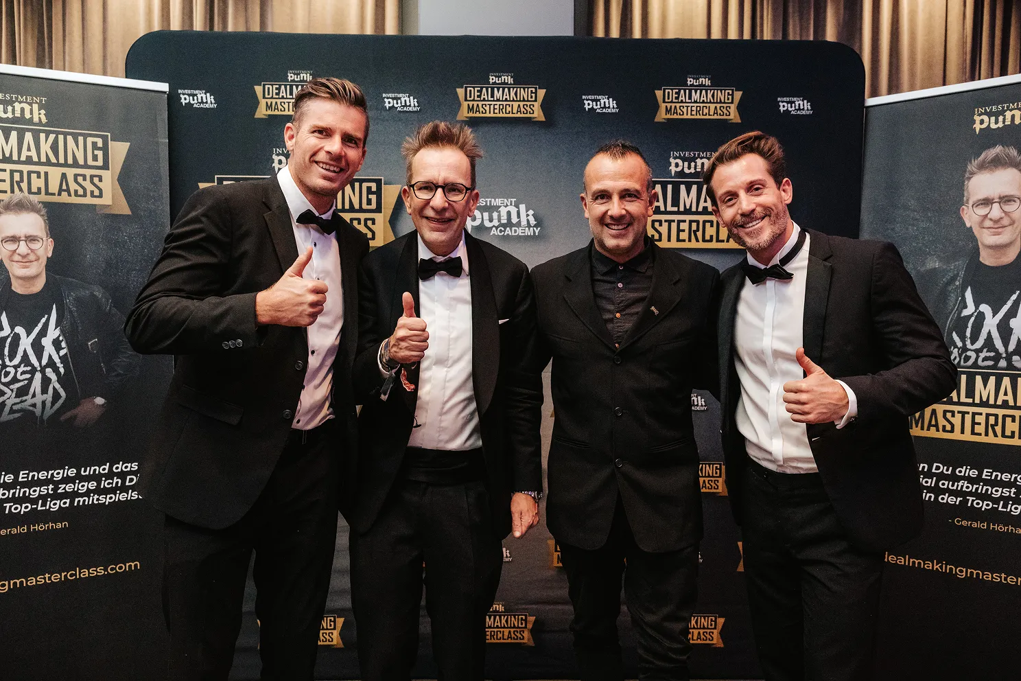 Vier Männer in formeller Kleidung posieren vor einem Banner der Investment Punk Dealmaking Masterclass, drei davon zeigen den Daumen hoch.