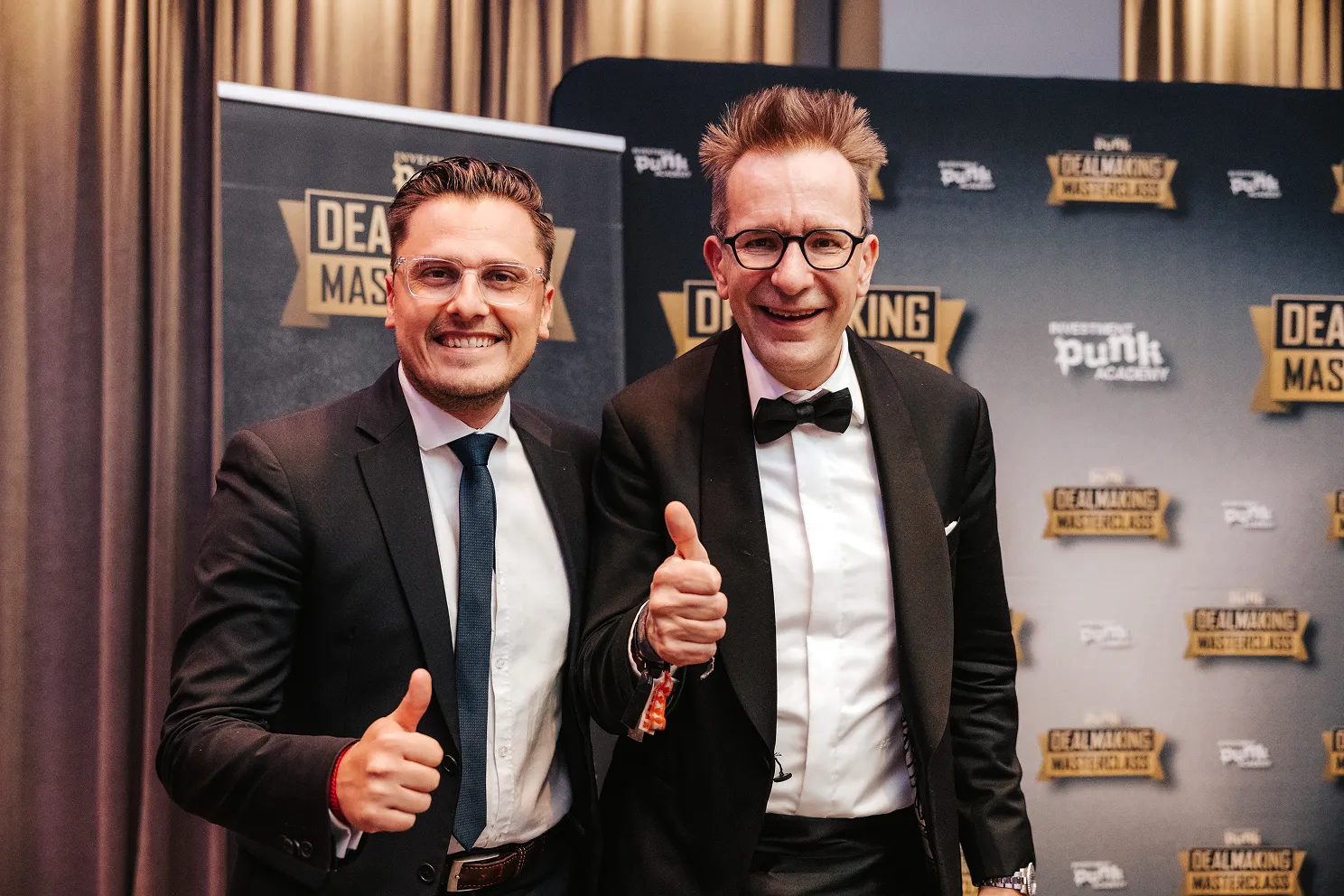 Zwei Männer in schwarzen Anzügen und Brillen lächeln und zeigen Daumen hoch vor einem Hintergrund mit 'Deal Making Masterclass' Bannern.