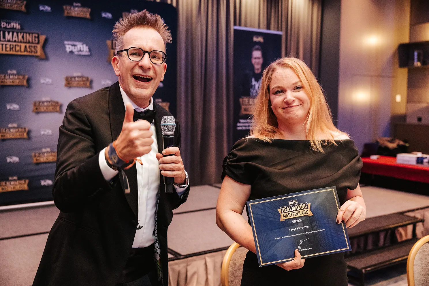 Ein Mann im schwarzen Anzug zeigt Daumen hoch und eine Frau hält eine Urkunde der Dealmaking Masterclass.