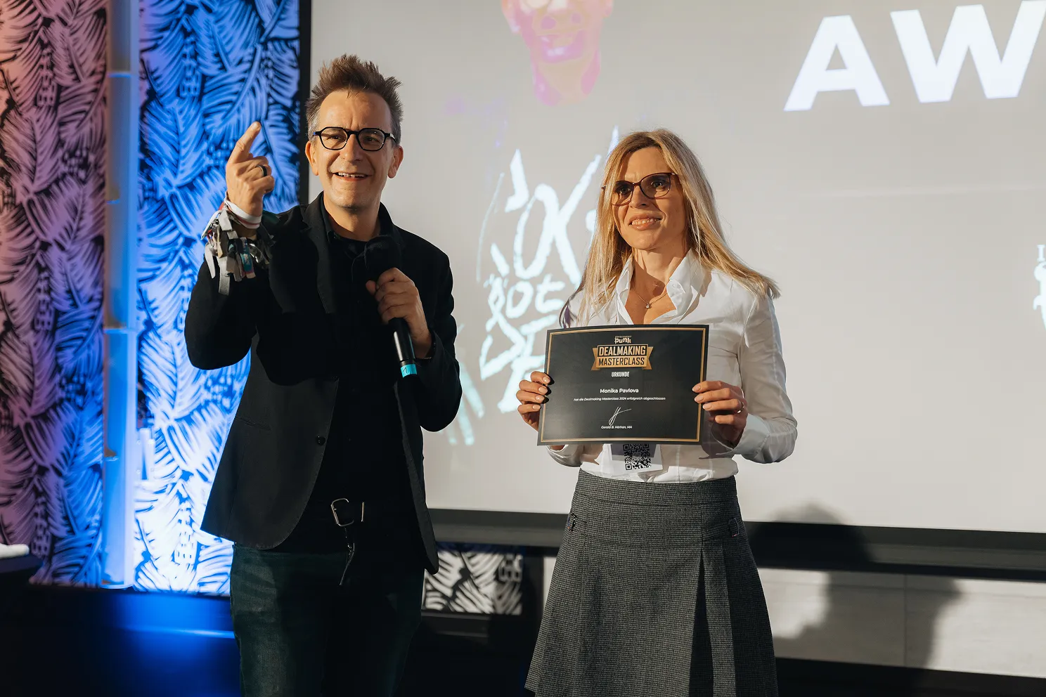 Zwei Personen stehen vor einer Leinwand, eine hält eine Urkunde mit dem Text 'Dealmaking Masterclass'.