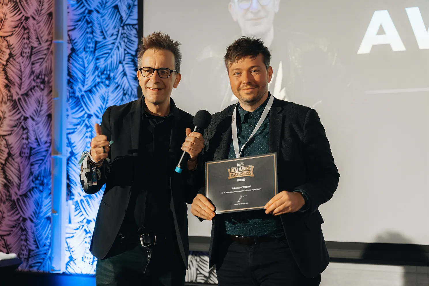 Zwei Männer stehen nebeneinander, einer hält ein Mikrofon und zeigt Daumen hoch, der andere hält eine Urkunde der Dealmaking Masterclass.