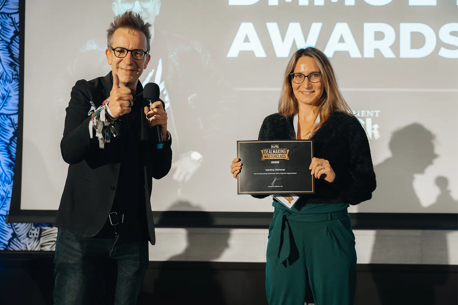 Eine Frau hält ein Zertifikat der Dealmaking Masterclass und ein Mann gibt einen Daumen hoch mit einem Mikrofon in der Hand.