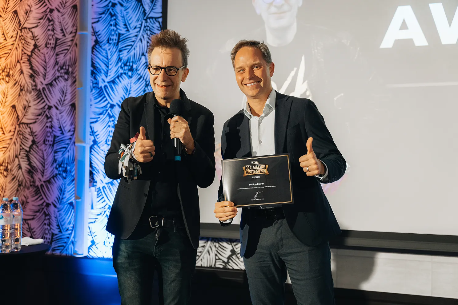 Zwei Männer in Anzügen lächeln und zeigen Daumen hoch, einer hält eine Urkunde der Dealmaking Masterclass.