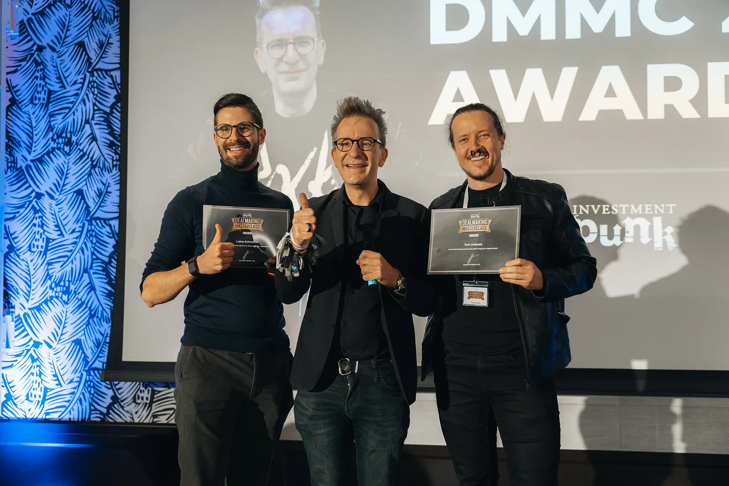 Drei Männer stehen vor einer Leinwand mit dem Schriftzug DMMC AWARD, zwei von ihnen halten Zertifikate der Dealmaking Masterclass und alle zeigen den Daumen hoch.