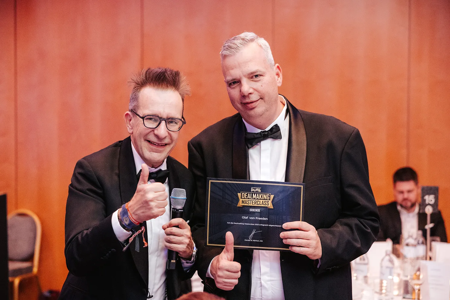 Zwei Männer in schwarzen Smokings, einer hält eine Urkunde und beide zeigen Daumen hoch.