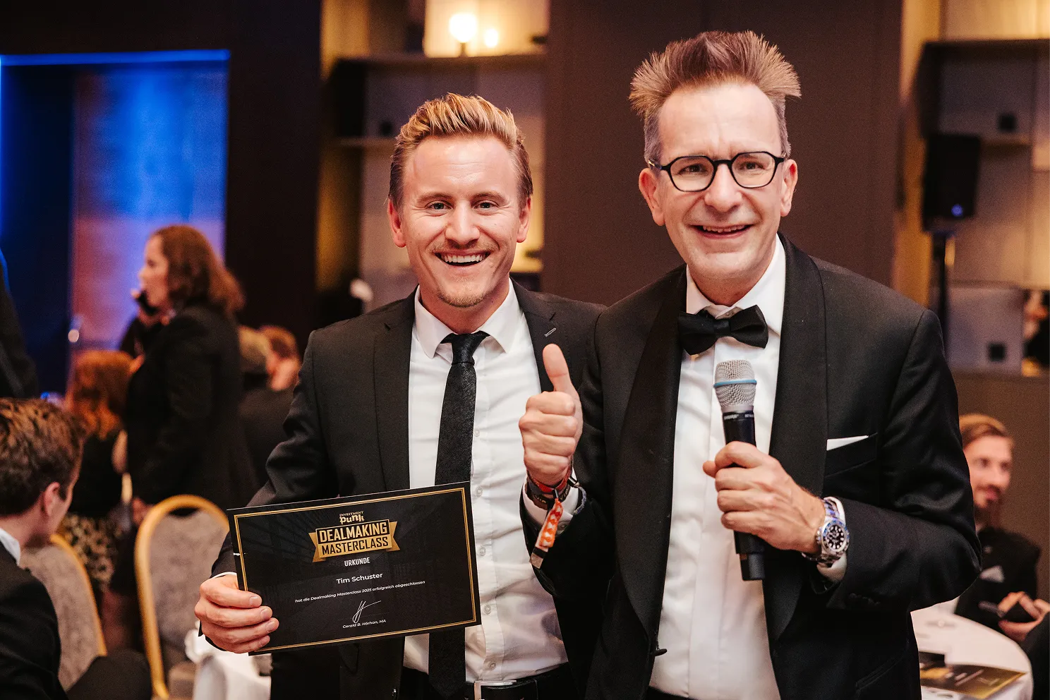 Zwei Männer in formeller Kleidung, einer hält ein Zertifikat der Dealmaking Masterclass und der andere ein Mikrofon und zeigt Daumen hoch.