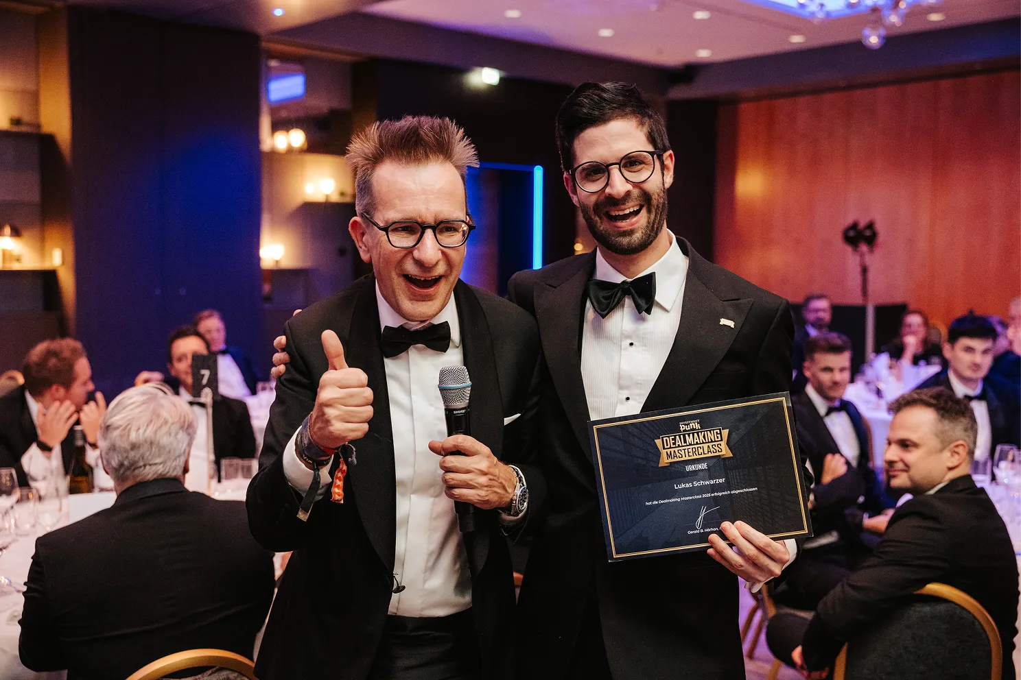 Zwei Männer in schwarzen Smokingjacken, einer mit Mikrofon gibt Daumen hoch, der andere hält eine Urkunde der Dealmaking Masterclass, während Gäste im Hintergrund sitzen.