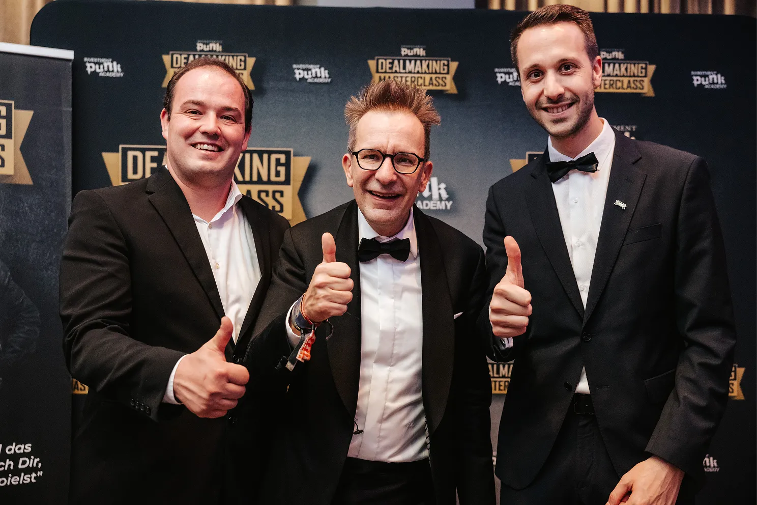 Drei Männer in schwarzen Anzügen und Fliegen zeigen lachend Daumen nach oben vor einem Banner mit der Aufschrift Dealmaking Masterclass.