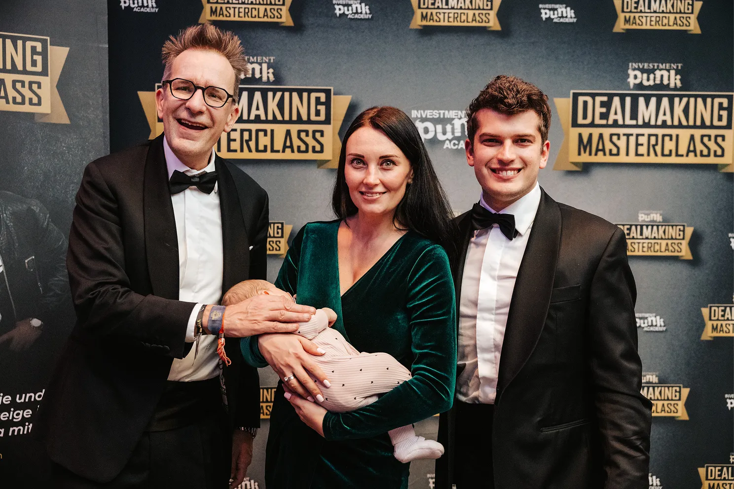 Zwei Männer in schwarzen Anzügen mit Fliegen und eine Frau im grünen Samtkleid, die ein schlafendes Baby hält, vor einem Hintergrund mit dem Schriftzug "Dealmaking Masterclass".