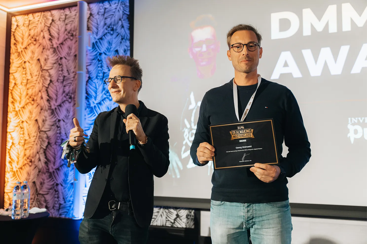 Zwei Männer bei der DMMC Veranstaltung, einer hält ein Mikrofon und zeigt Daumen hoch, der andere hält eine Urkunde der Dealmaking Masterclass.