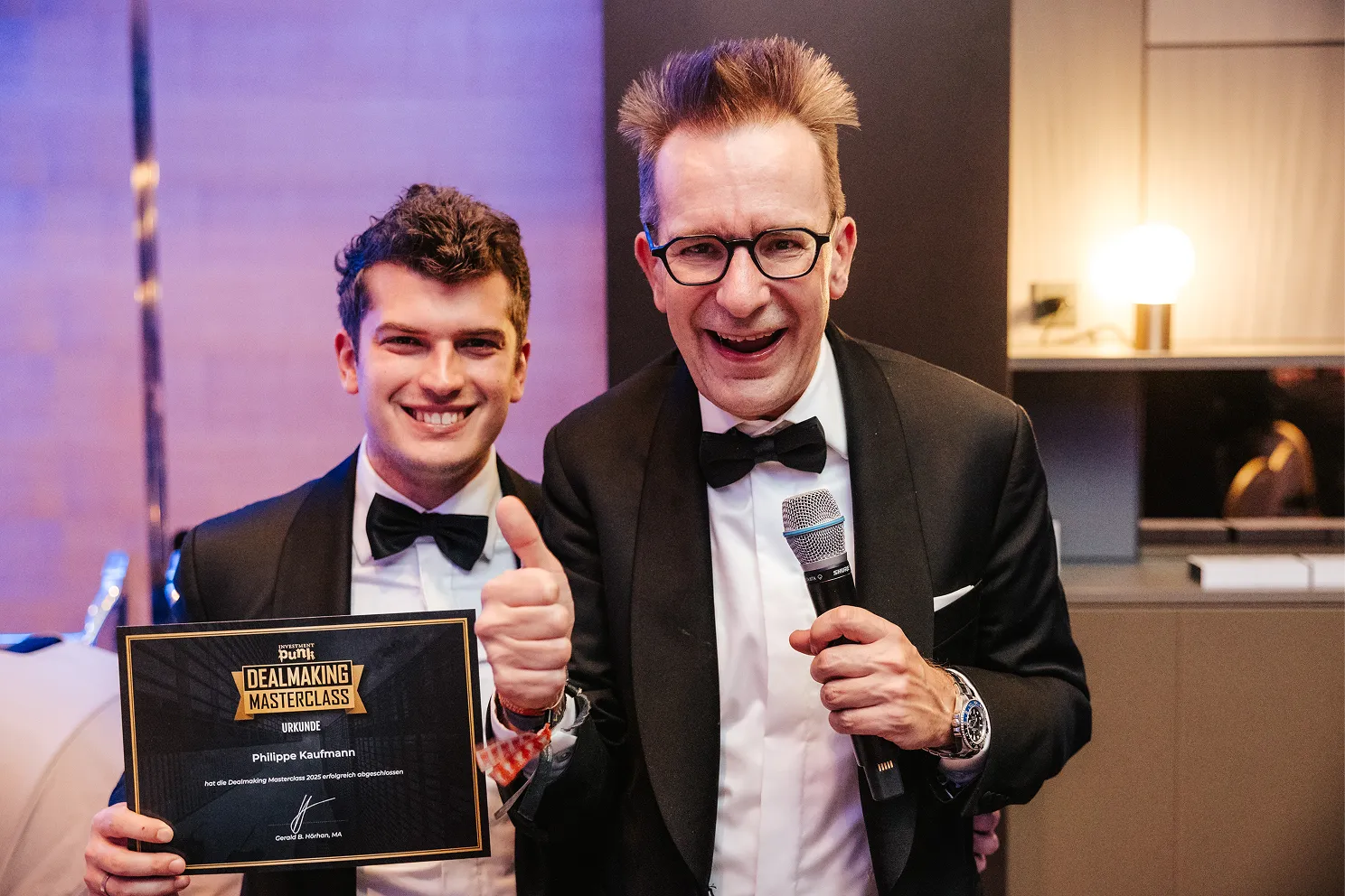 Zwei Männer in schwarzer Abendkleidung, einer hält eine Urkunde vom Dealmaking Masterclass 2025, der andere zeigt Daumen hoch und hält ein Mikrofon.
