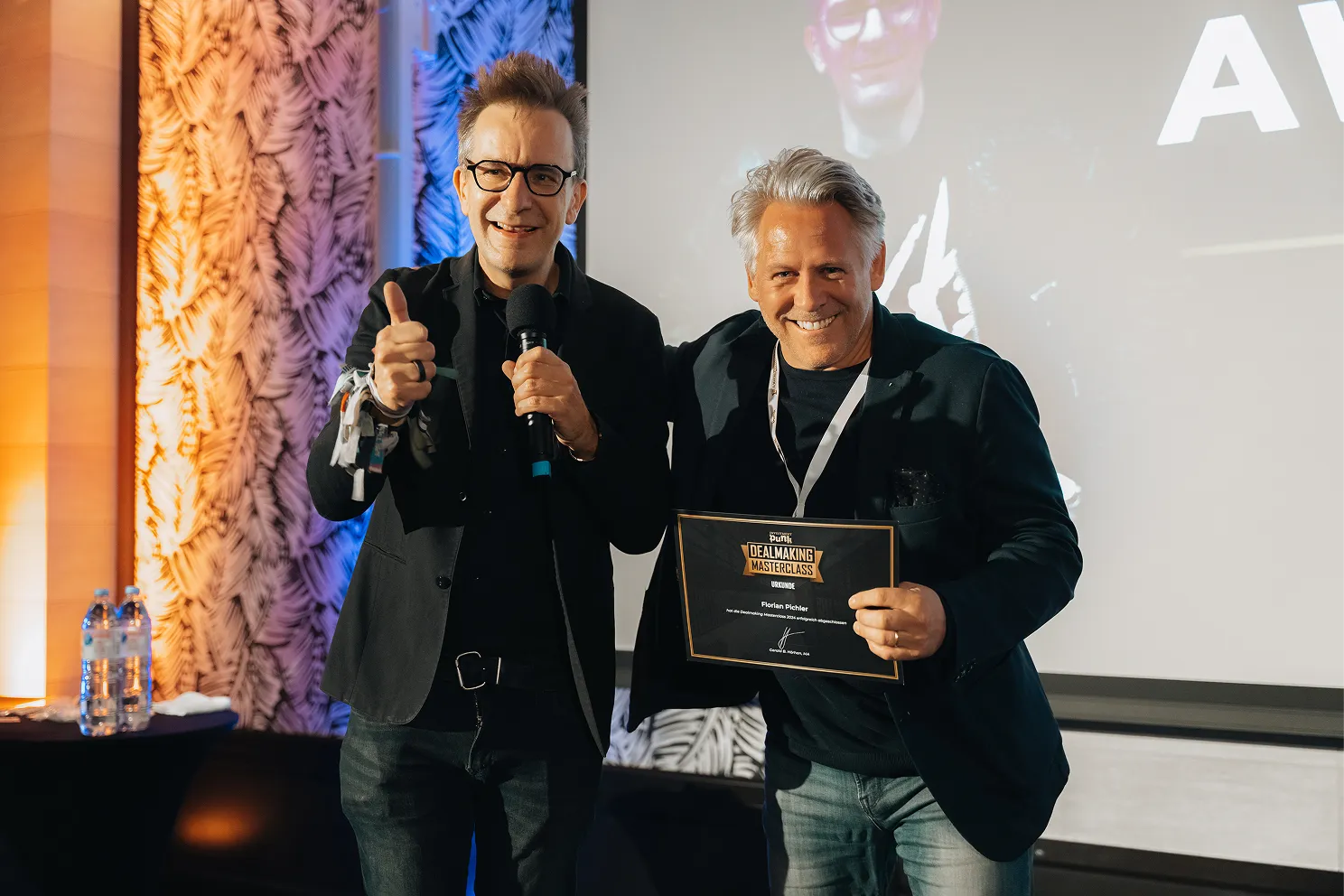 Zwei lächelnde Männer, einer mit Mikrofon zeigt Daumen hoch, der andere hält eine Urkunde der DealMaking Masterclass.