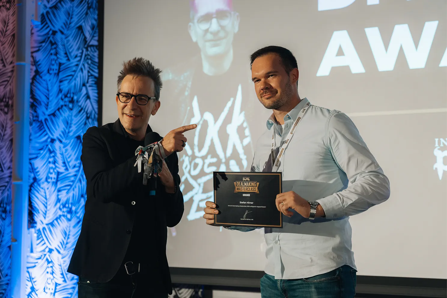 Zwei Männer auf einer Bühne, einer hält eine Urkunde mit der Aufschrift 'Dealmaking Masterclass' und der andere zeigt darauf.