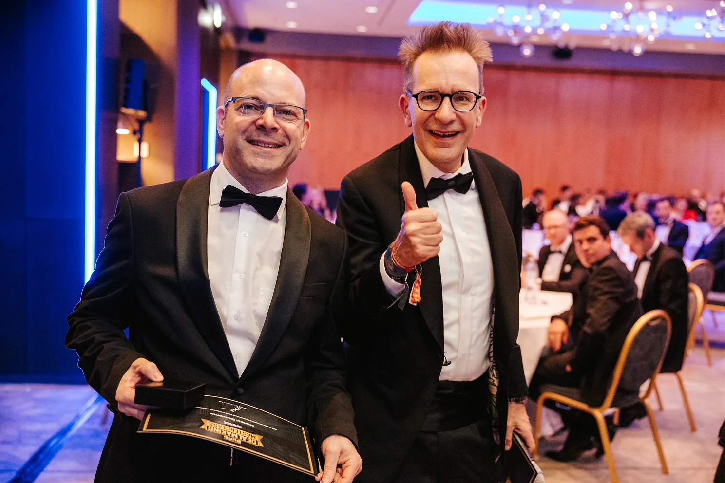 Zwei Männer im Smoking bei einer festlichen Veranstaltung, einer hält eine Urkunde und ein Kästchen, der andere zeigt Daumen hoch.