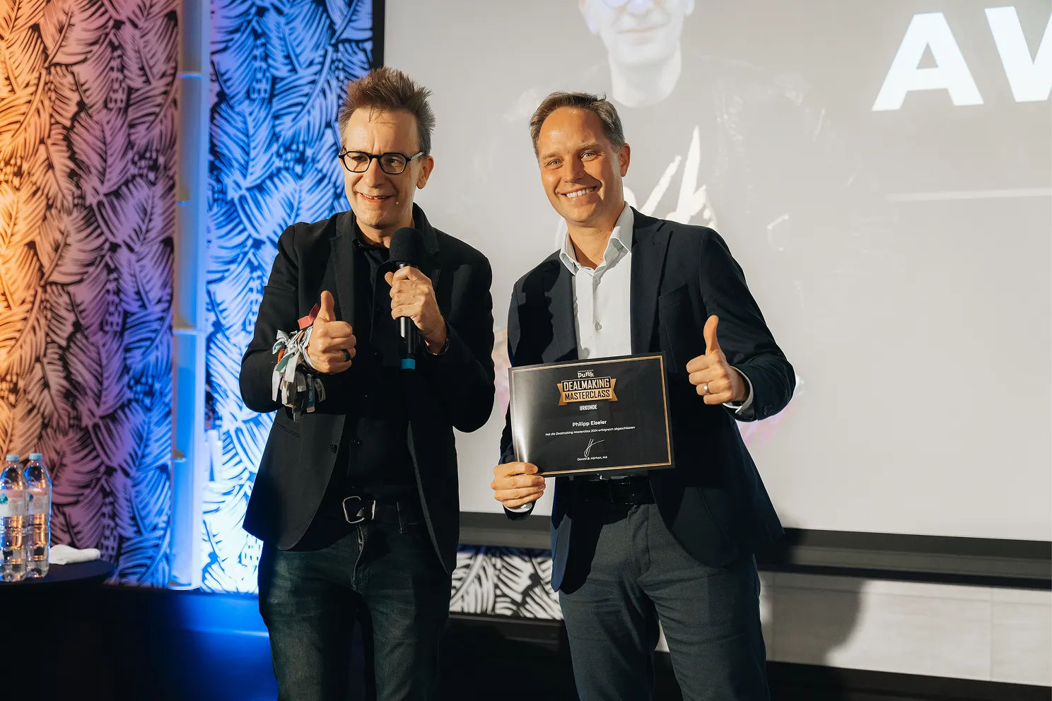 Zwei Männer in Anzügen lächeln und zeigen Daumen hoch, einer hält eine Dealmaking Masterclass Urkunde.