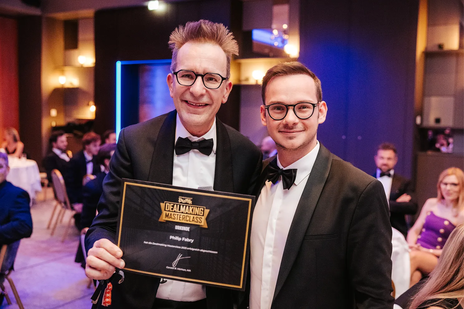 Zwei Männer in schwarzen Anzügen mit Fliegen, einer hält eine Urkunde der Punk Dealmaking Masterclass.