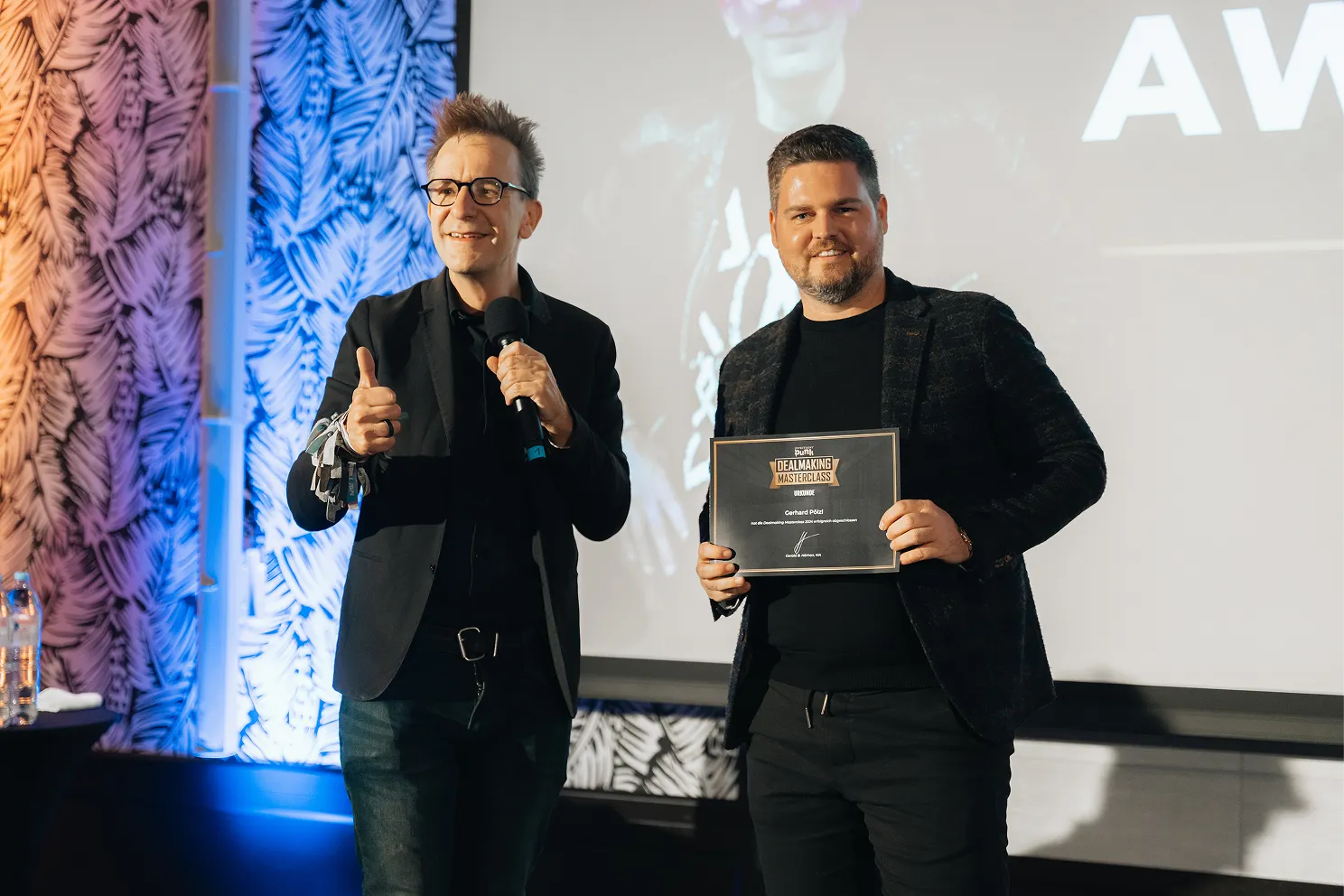 Zwei Männer auf einer Bühne, einer hält ein Mikrofon und zeigt Daumen hoch, der andere hält ein Zertifikat für die Dealmaking Masterclass.