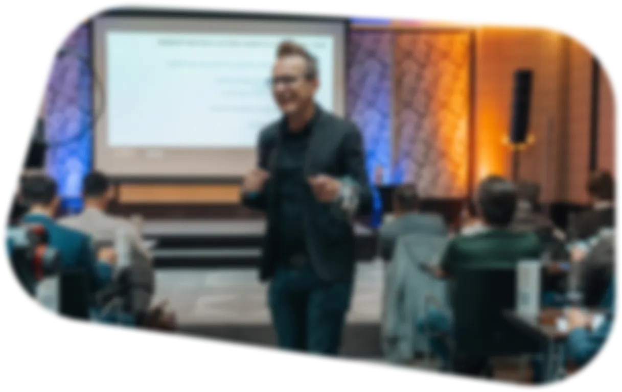 Ein verschwommener Vortragender steht vor einem Publikum in einem Konferenzraum mit einer Präsentation auf einer Leinwand.