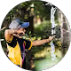 Man aiming a compound bow in a curved shooting stance outdoors, wearing a yellow shirt and a cap, surrounded by greenery.
