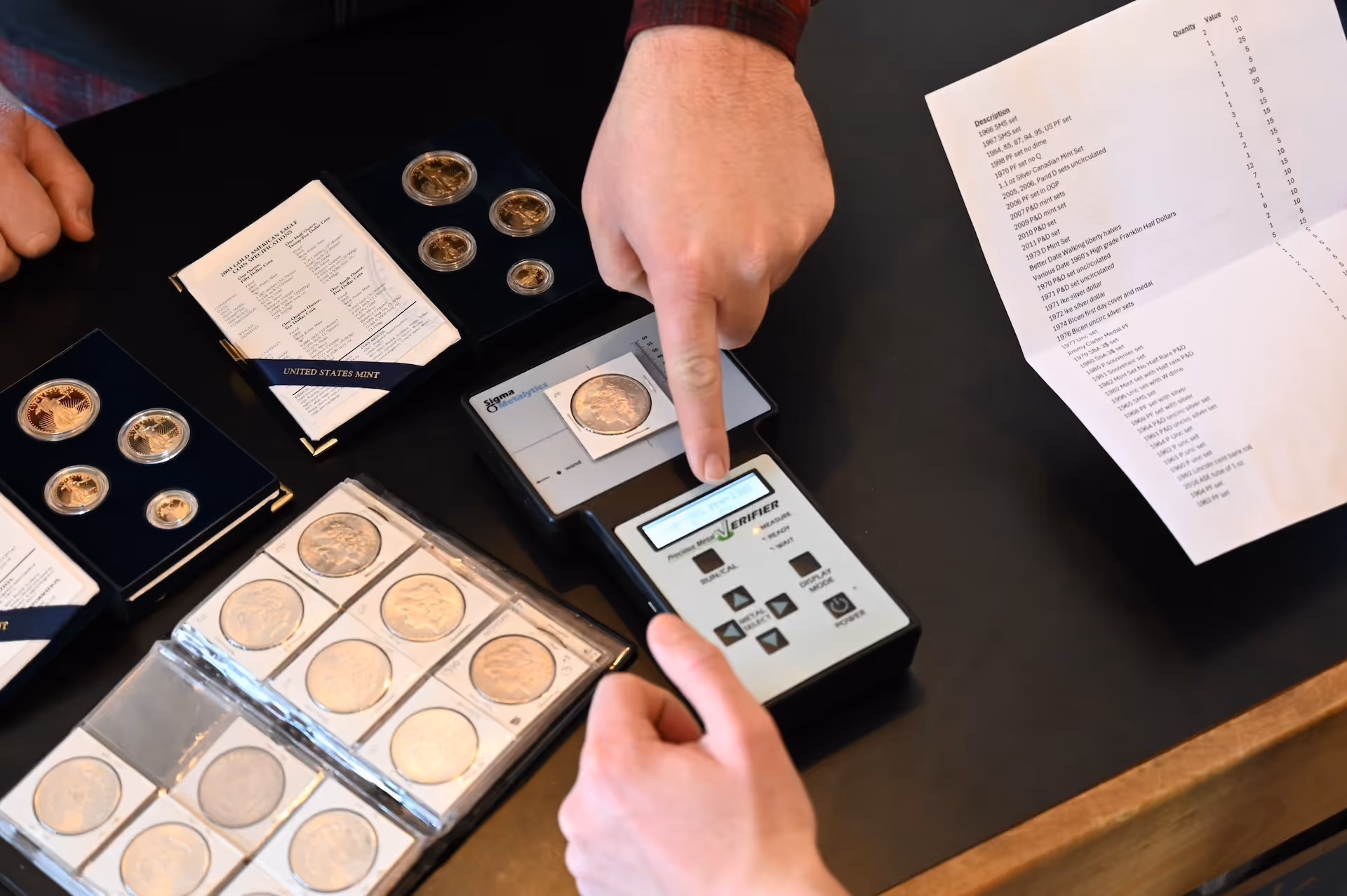 Two hands using a Verifier device to authenticate a coin placed on the machine, surrounded by coin collections and a list on a table.