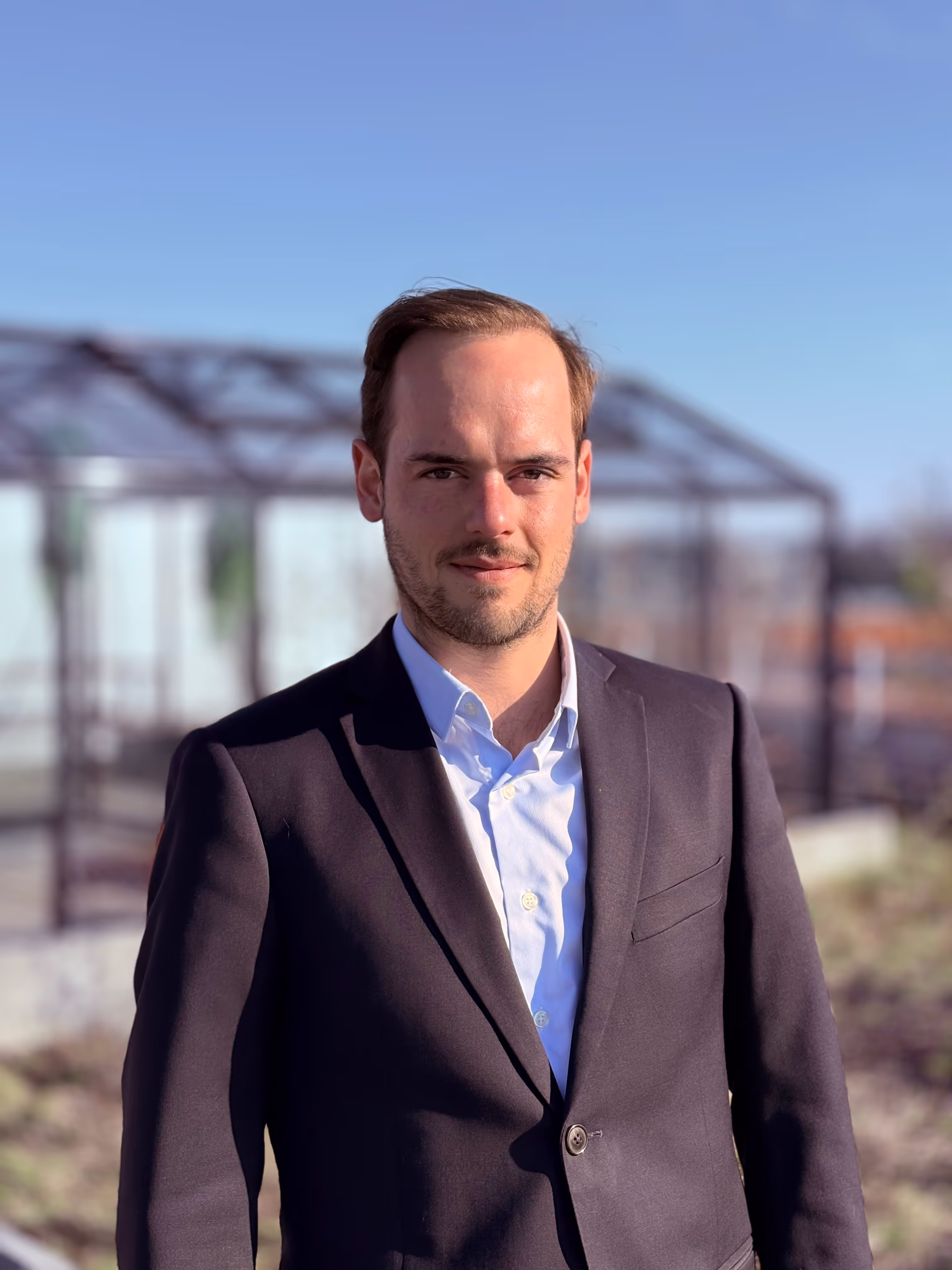 Eloi Ducrocq, fondateur de DUNE Energie en costume noir et chemise bleue devant une serre sous un ciel bleu clair.