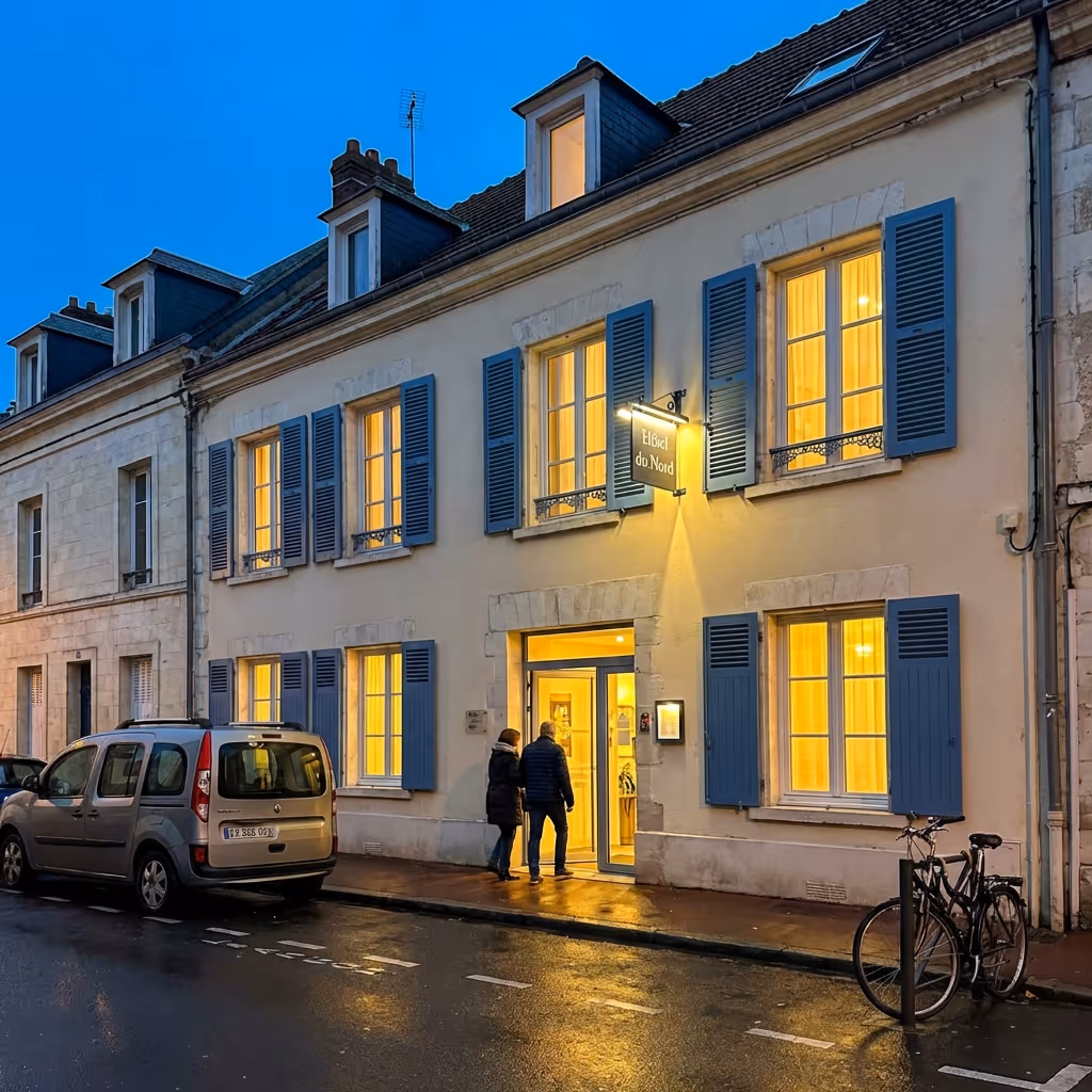 Façade d'un hôtel client DUNE Energie, avec volets bleus, lumière chaude aux fenêtres, une voiture et deux personnes à l'entrée.