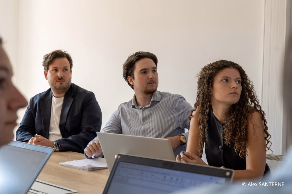 Trois jeunes adultes assis à une table lors d'une réunion, avec des ordinateurs portables ouverts.
