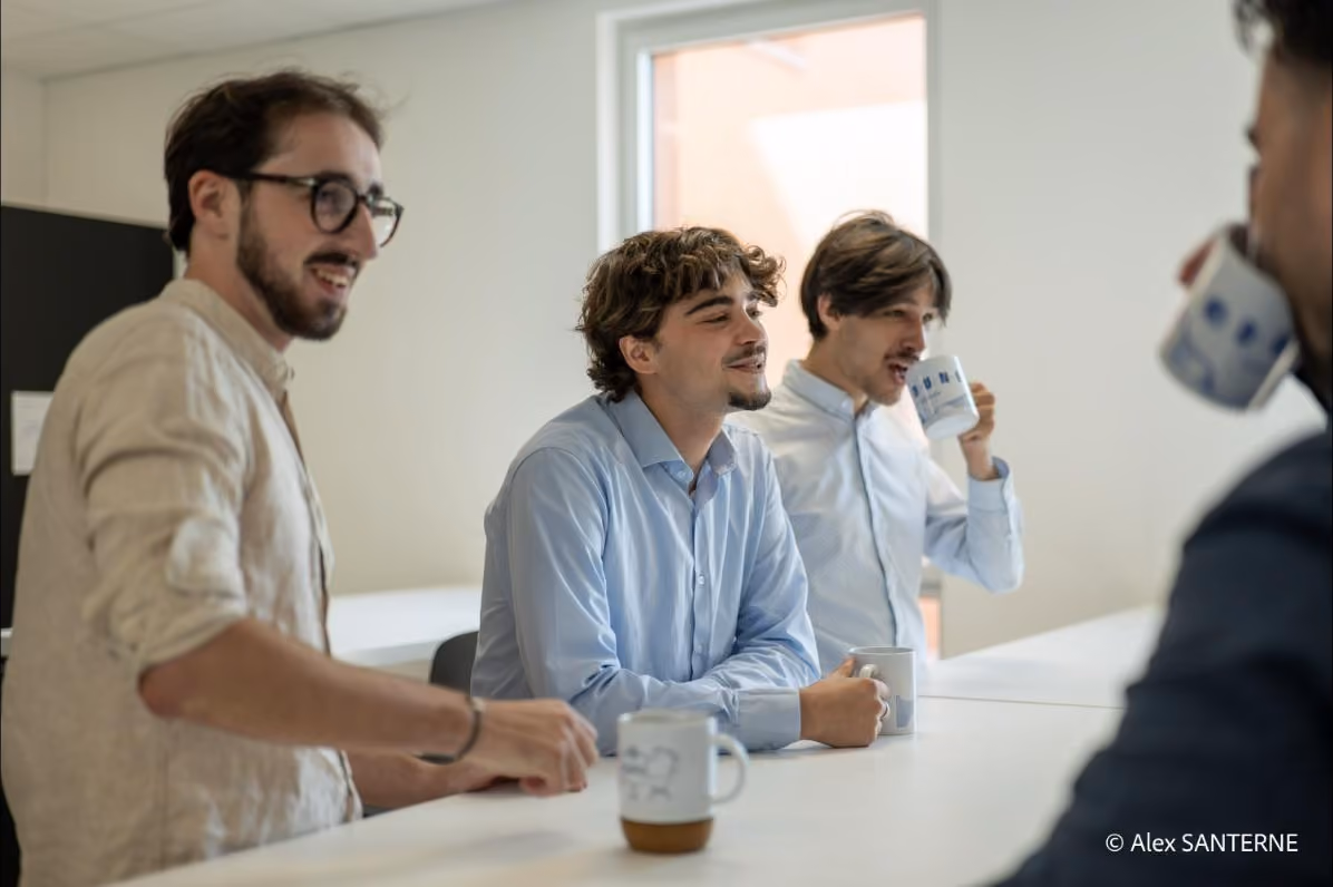 Quatre hommes bavardant et buvant du café autour d'une table dans une pièce lumineuse.
