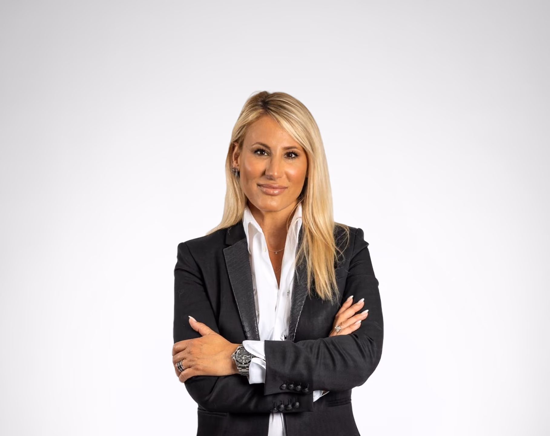 Femme blonde en costume noir, chemise blanche, bras croisés, posant devant fond gris clair.