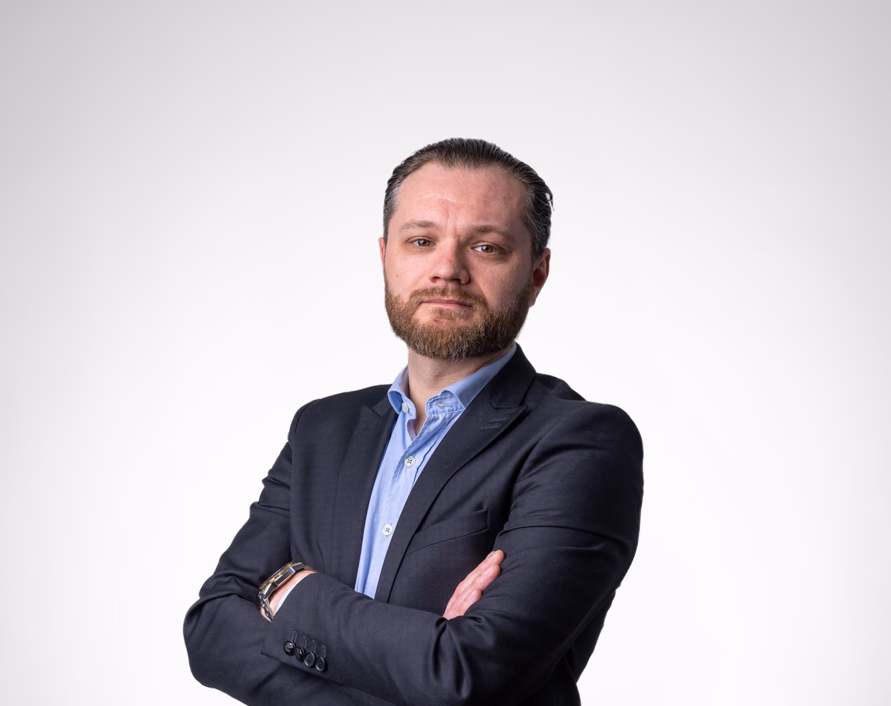 Homme d'affaires avec barbe, chemise bleue et costume sombre, bras croisés devant un fond clair.