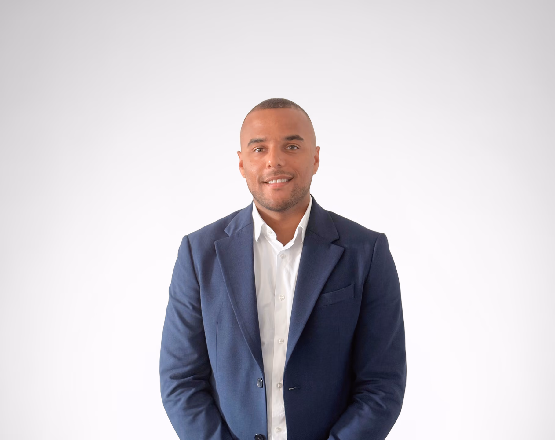 Homme souriant en veste bleu marine et chemise blanche posant devant un mur blanc.